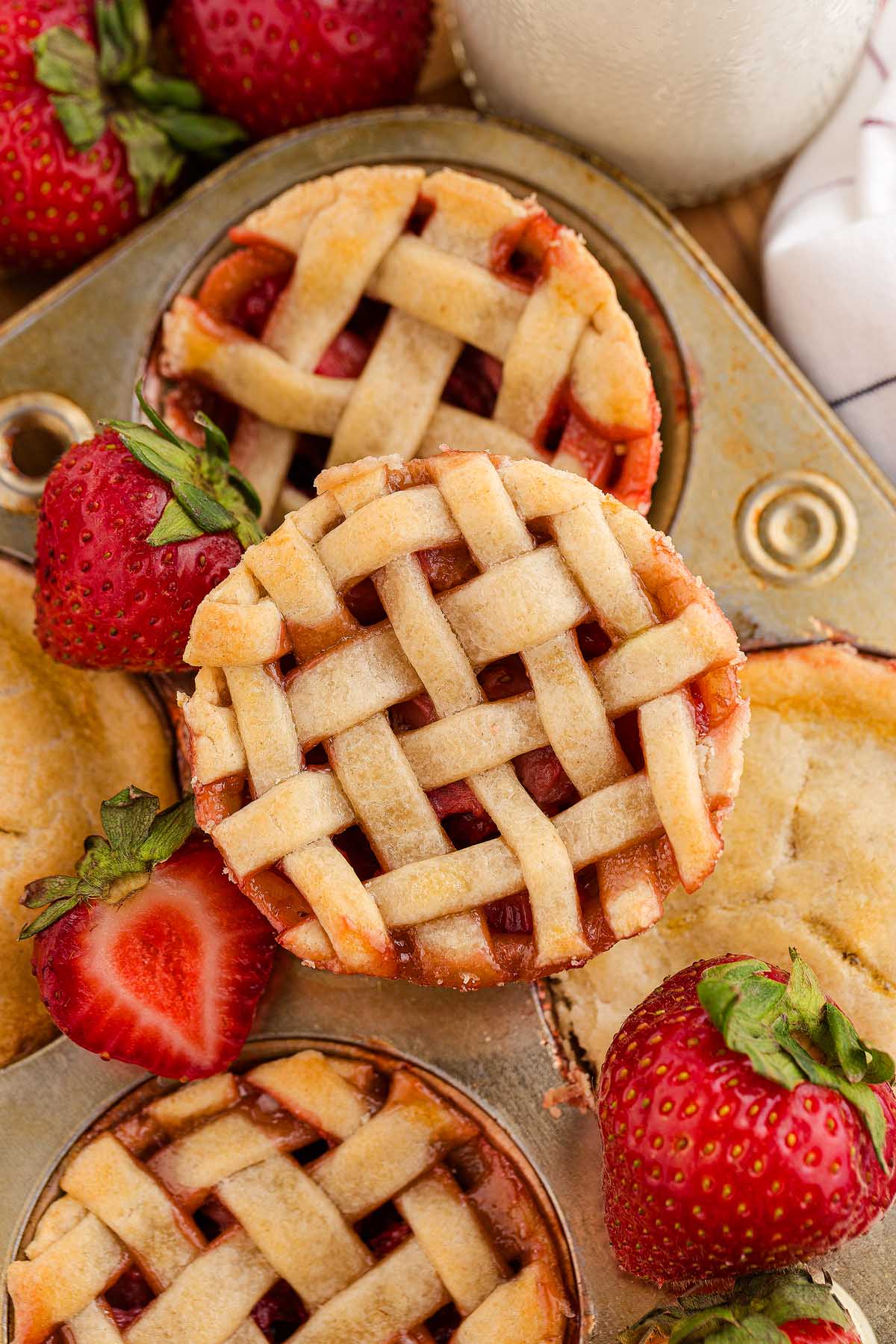 Mini strawberry pies with lattice crusts baked in a muffin tin, surrounded by fresh strawberries, make a charming and delicious treat.