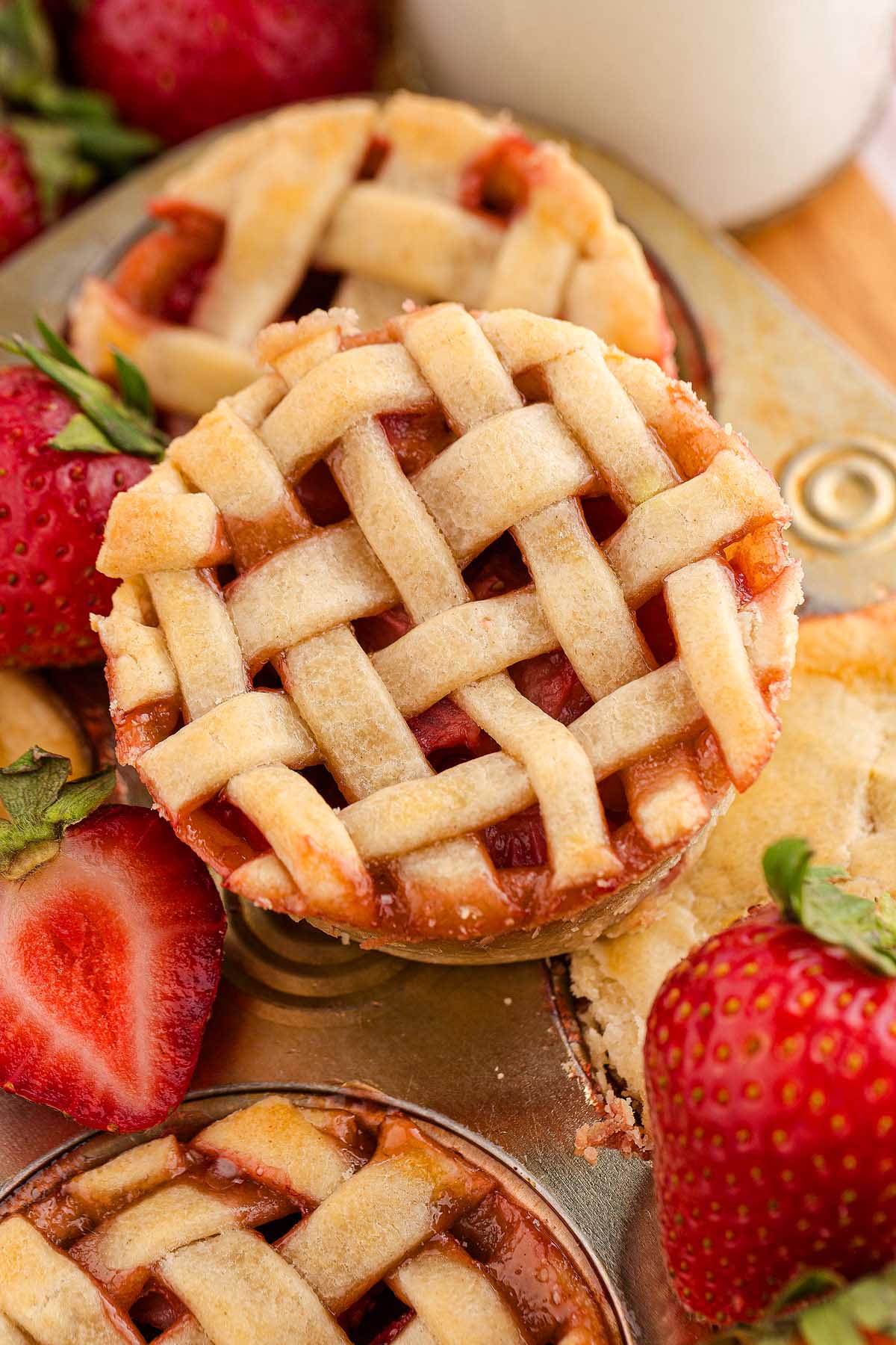 Mini strawberry pies with delicate lattice crusts are baked in a muffin tin, surrounded by fresh, juicy strawberries.