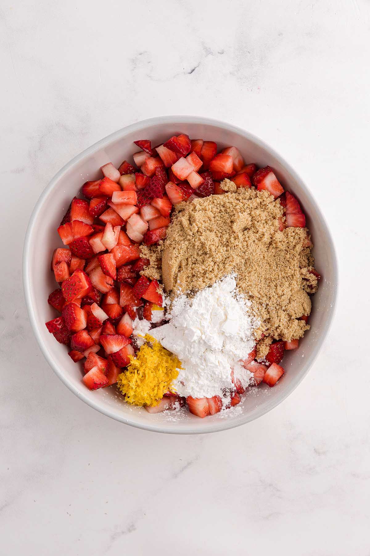 Chopped strawberries, brown sugar, cornstarch, and lemon zest in a white bowl on a white surface—perfect for filling mini strawberry pies.