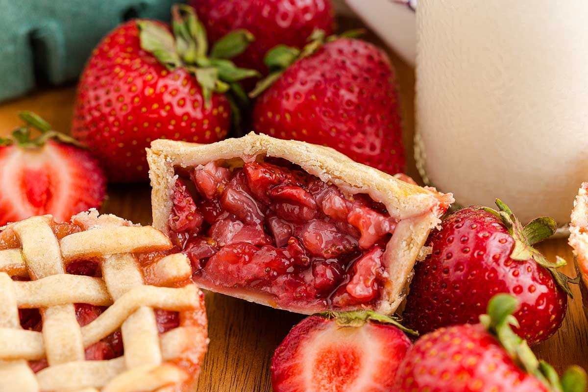 A mini strawberry pie with a juicy filling sits among fresh strawberries and a glass of milk, showcasing the irresistible charm of mini strawberry pies.