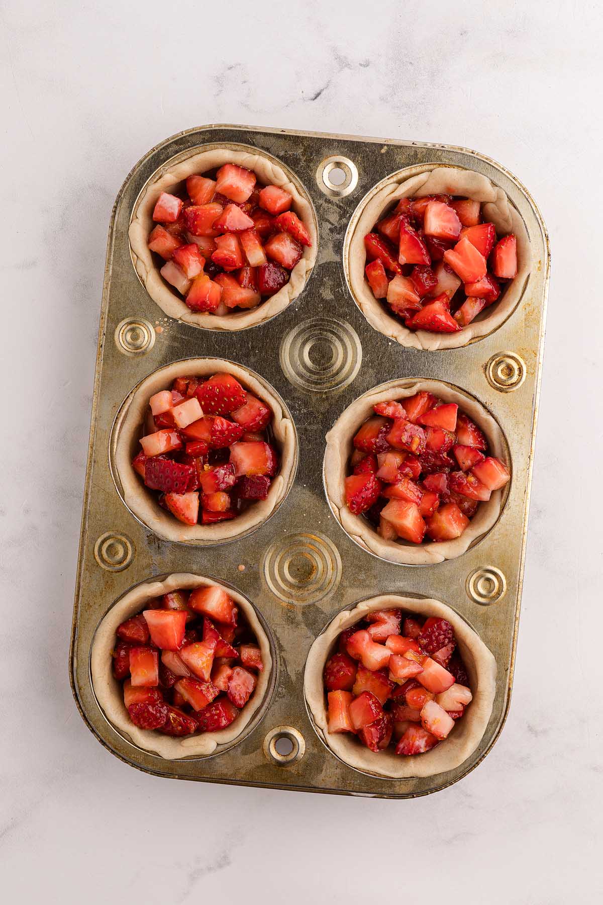 A muffin tin with six unbaked mini strawberry pies filled with chopped strawberries rests on a white surface.