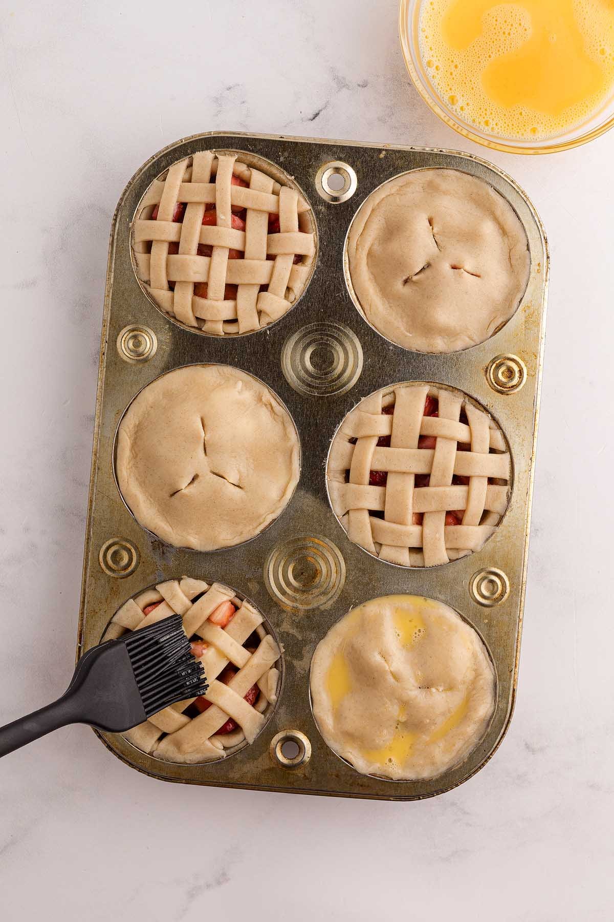 A muffin tin with six unbaked mini strawberry pies, some topped with lattice crusts, being brushed with egg wash.
