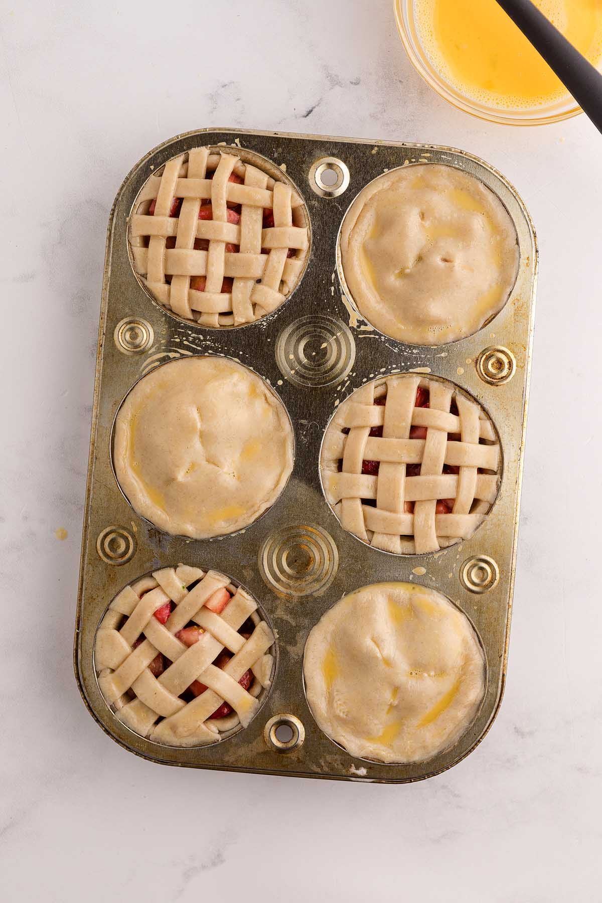 A muffin tin filled with six unbaked mini strawberry pies, some topped with lattice crusts and others with solid crusts.