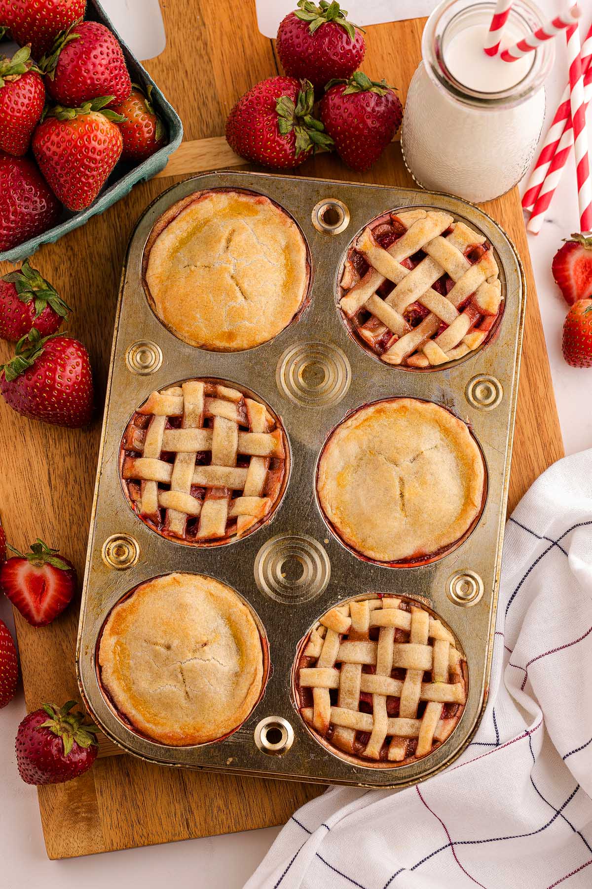 A muffin tin filled with mini strawberry pies, fresh strawberries, and a bottle of milk with straws sit on a wooden board.