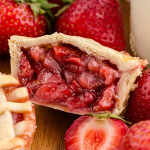 A mini strawberry pie cut in half, surrounded by fresh strawberries and another of our delicious mini strawberry pies.