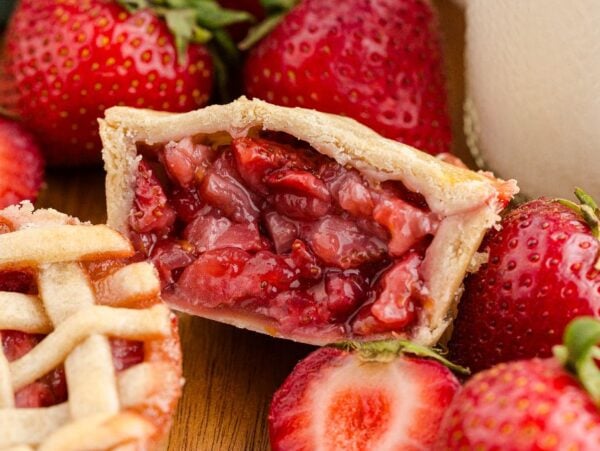A mini strawberry pie cut in half, surrounded by fresh strawberries and another of our delicious mini strawberry pies.
