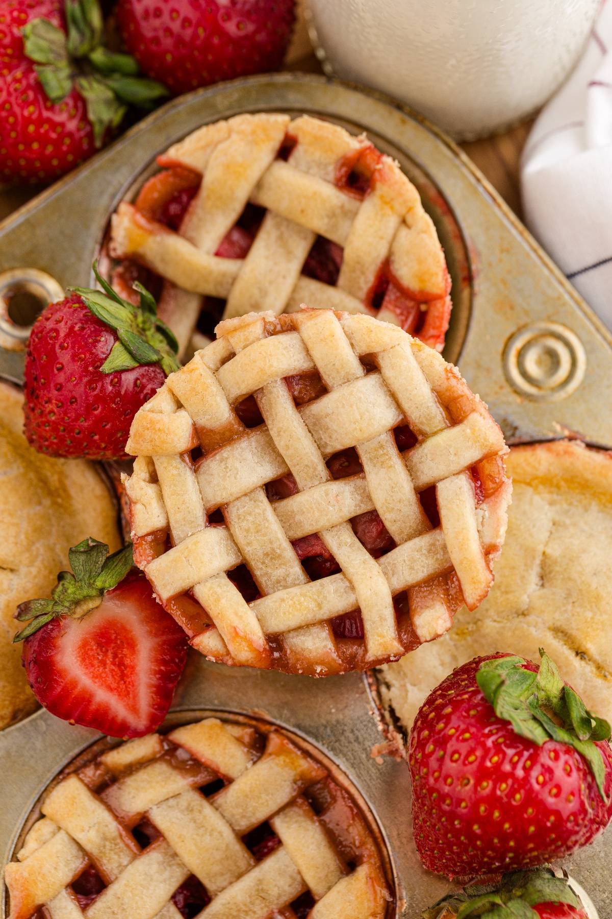 Mini strawberry pies with lattice crusts in a muffin tin, surrounded by fresh strawberries.