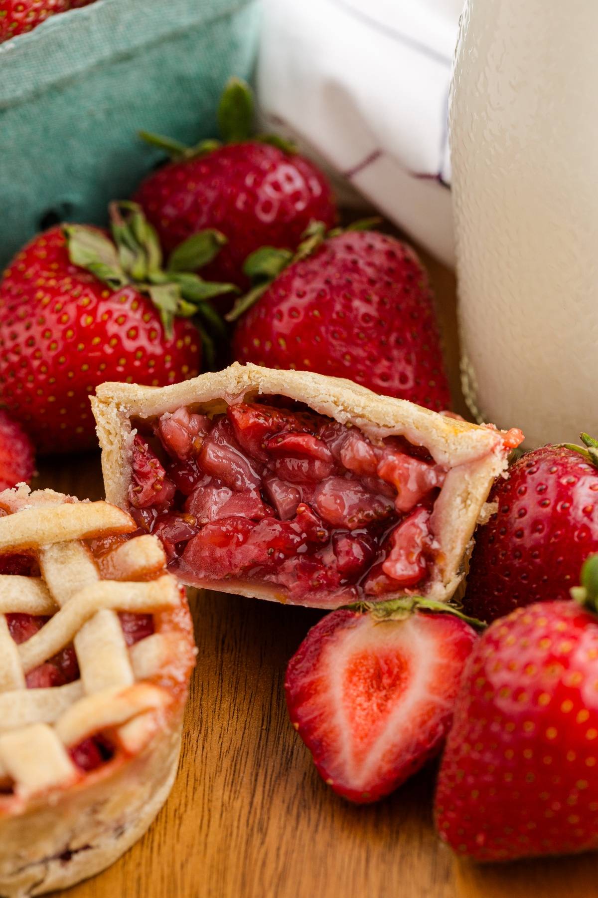 A mini strawberry pie cut open, showing the filling, surrounded by fresh strawberries on a wooden surface.