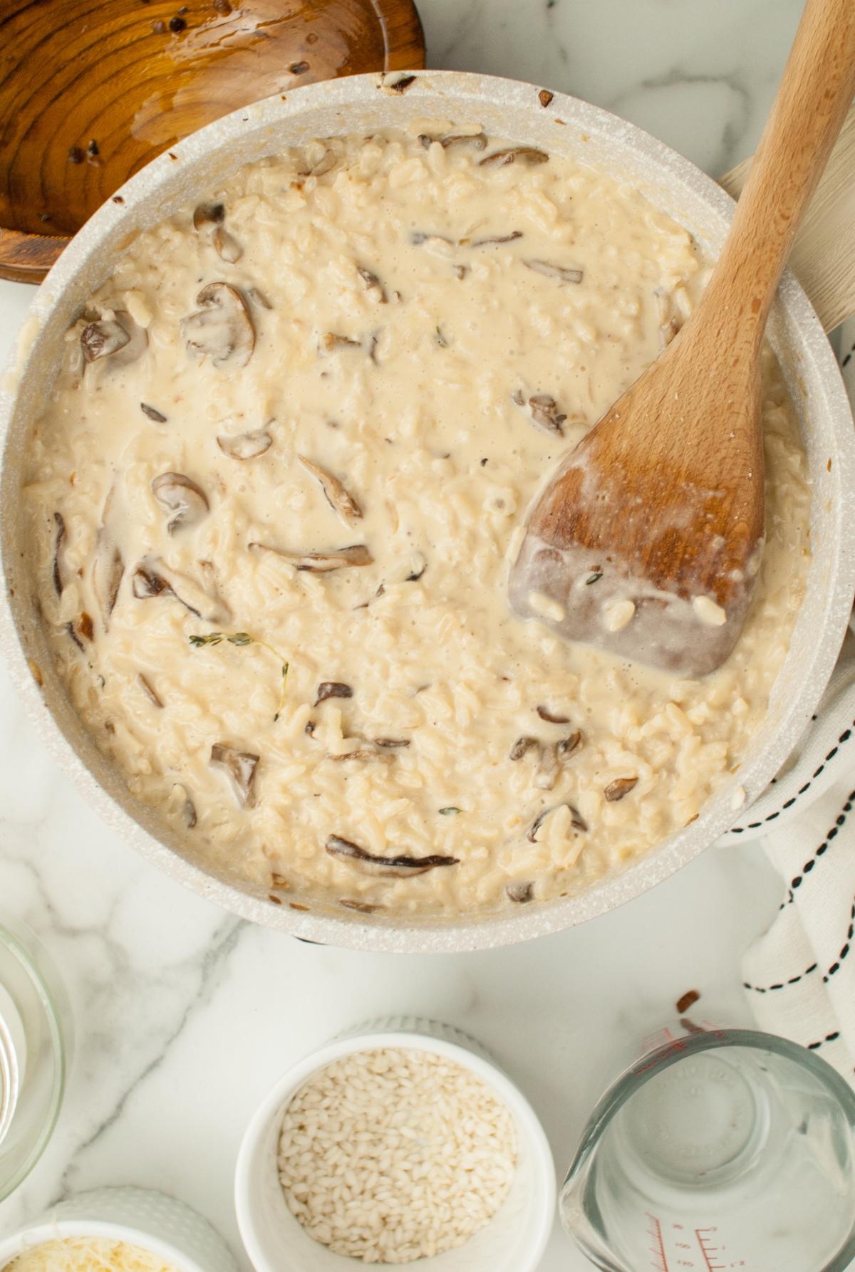 Creamy mushroom risotto featuring shiitake and cremini (baby bella) mushrooms is stirred in a pan with a wooden spoon on a marble countertop—an elegant side dish for chicken or turkey.
