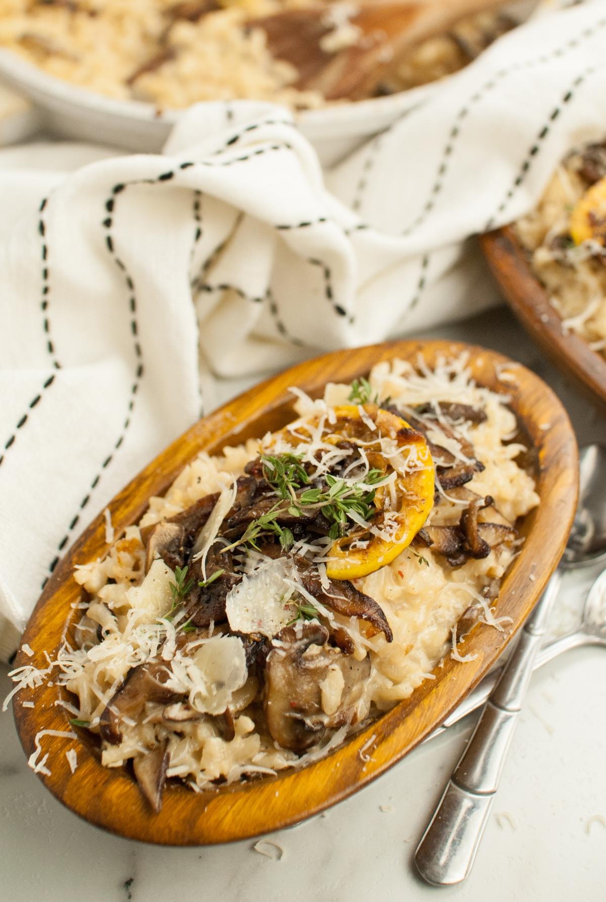 Oval wooden bowl filled with creamy mushroom risotto made with shiitake and cremini (baby bella) mushrooms, topped with herbs and grated cheese, beside a striped napkin.