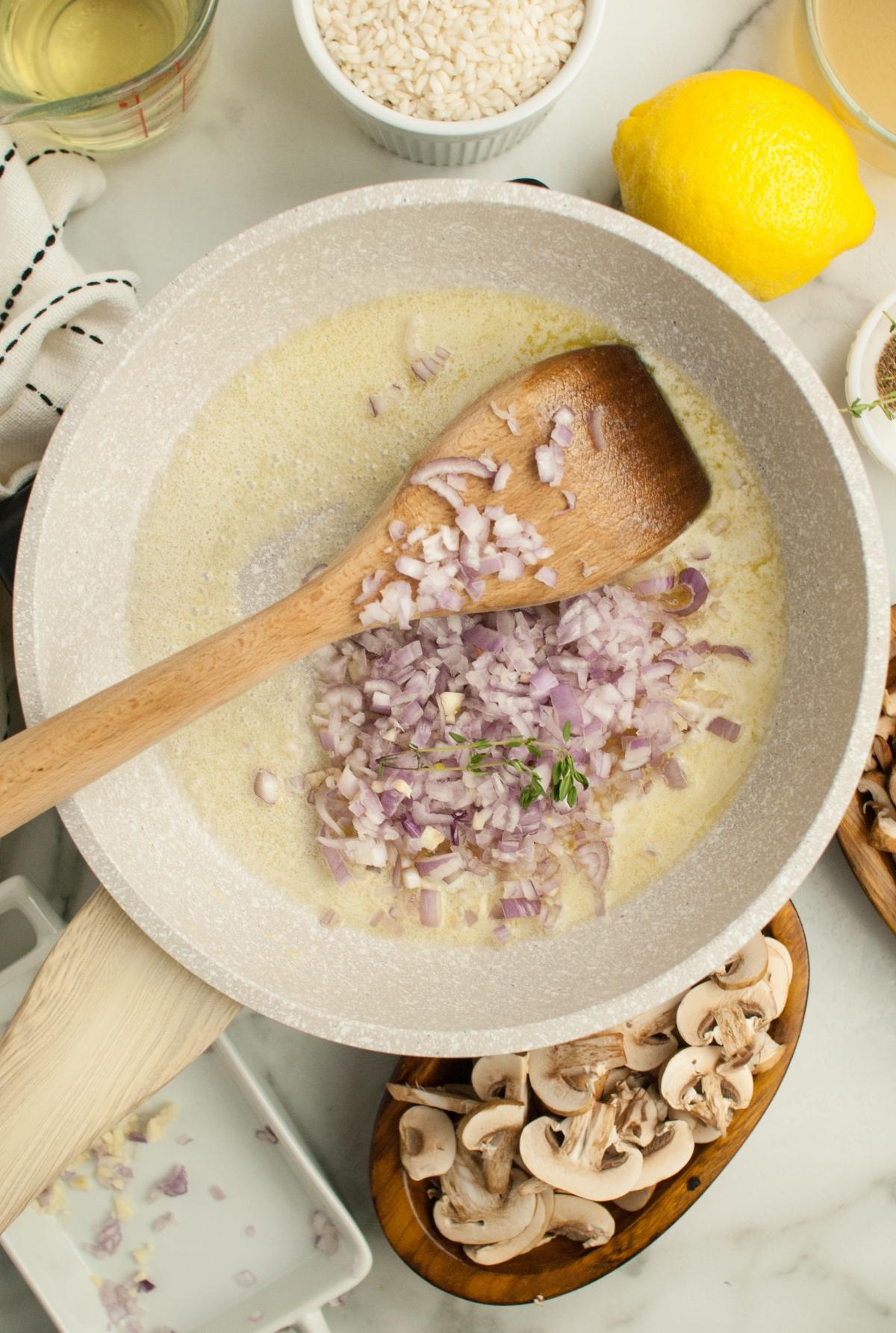 Chopped shallots cooking in a pan with butter, stirred with a wooden spoon; mushrooms and ingredients nearby for a simple risotto recipe—perfect as a side dish for chicken or turkey.