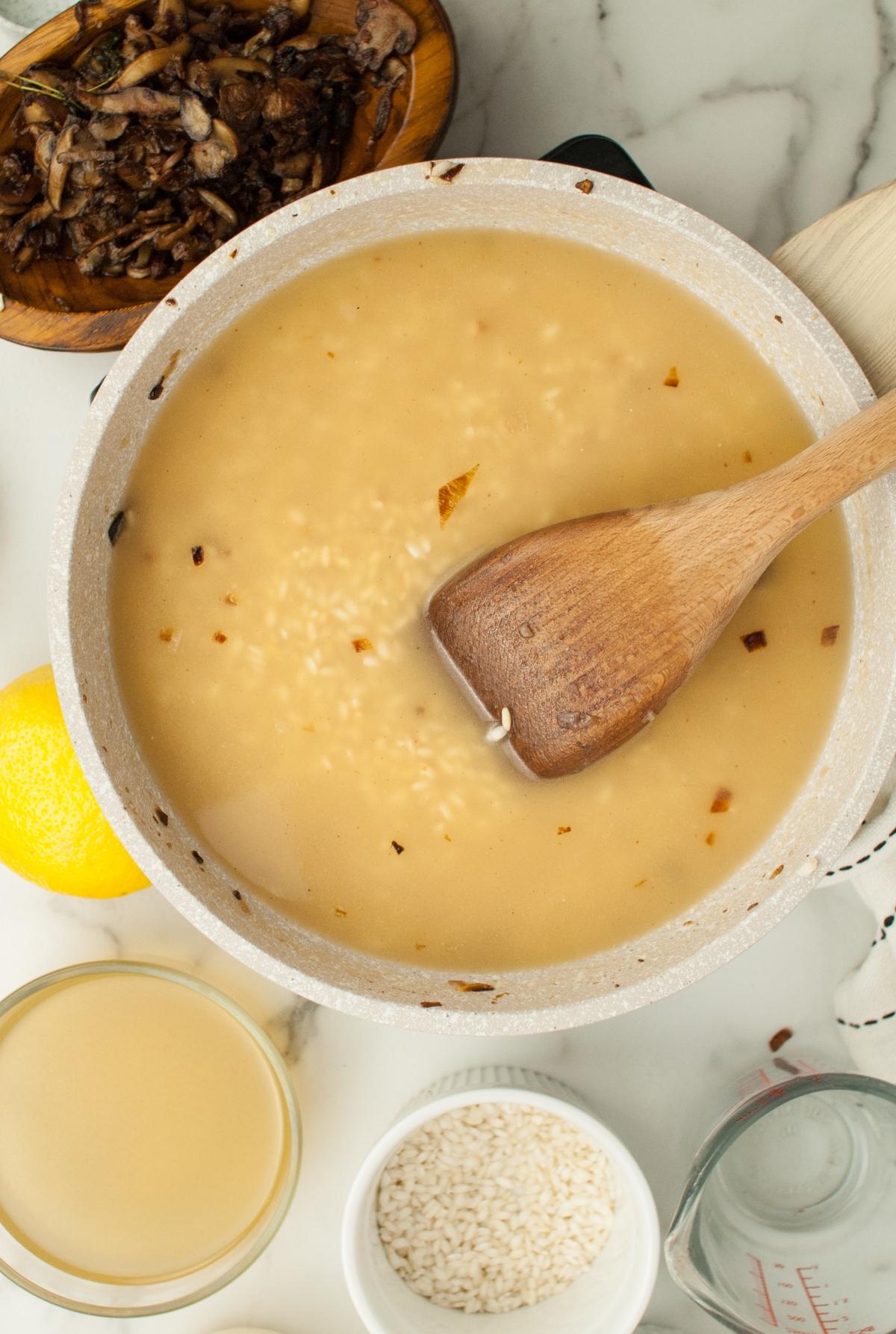 A pot of mushroom risotto being stirred with a wooden spoon, surrounded by fresh ingredients and a glass of Chardonnay on a marble countertop—a simple risotto recipe perfect for any home cook.