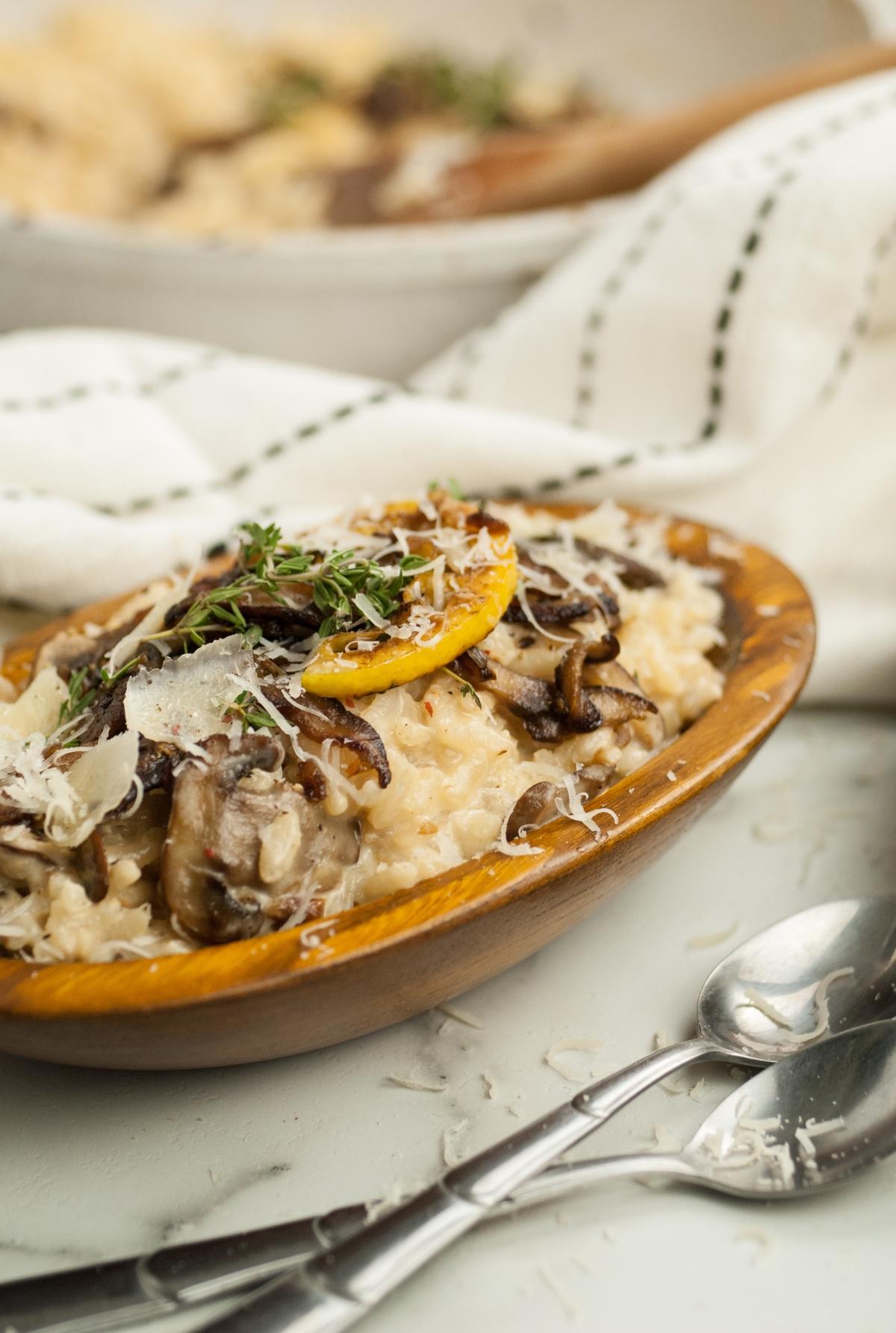 A wooden bowl of creamy mushroom risotto made with shiitake and cremini (baby bella), topped with herbs and grated cheese, sits next to spoons and a napkin.