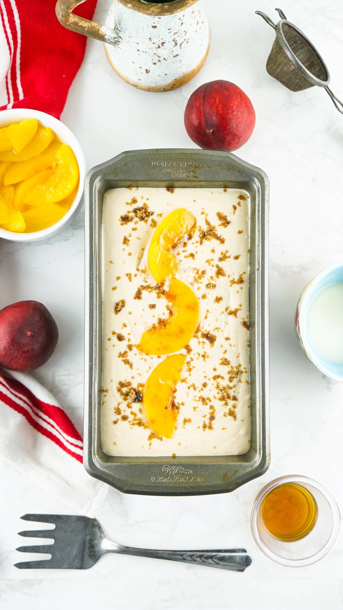 A loaf pan with batter topped with peach slices and crumbs, surrounded by peaches, syrup, and kitchen items—perfect for desserts made with canned peaches.
