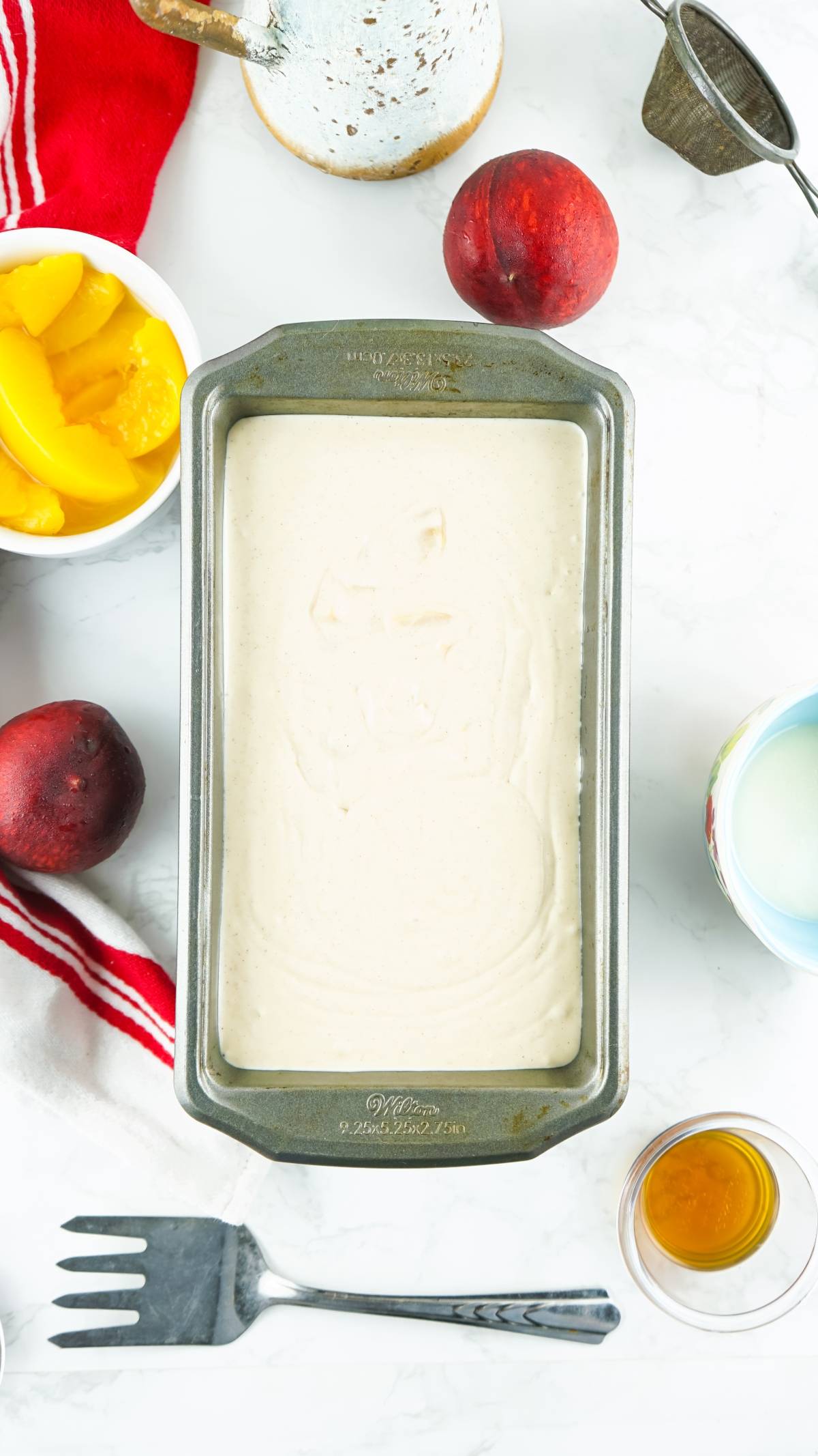 Loaf pan with cake batter, surrounded by peaches, milk, vanilla, and baking utensils on a white surface—perfect for crafting a boozy peach dessert or a treat made with canned peaches.