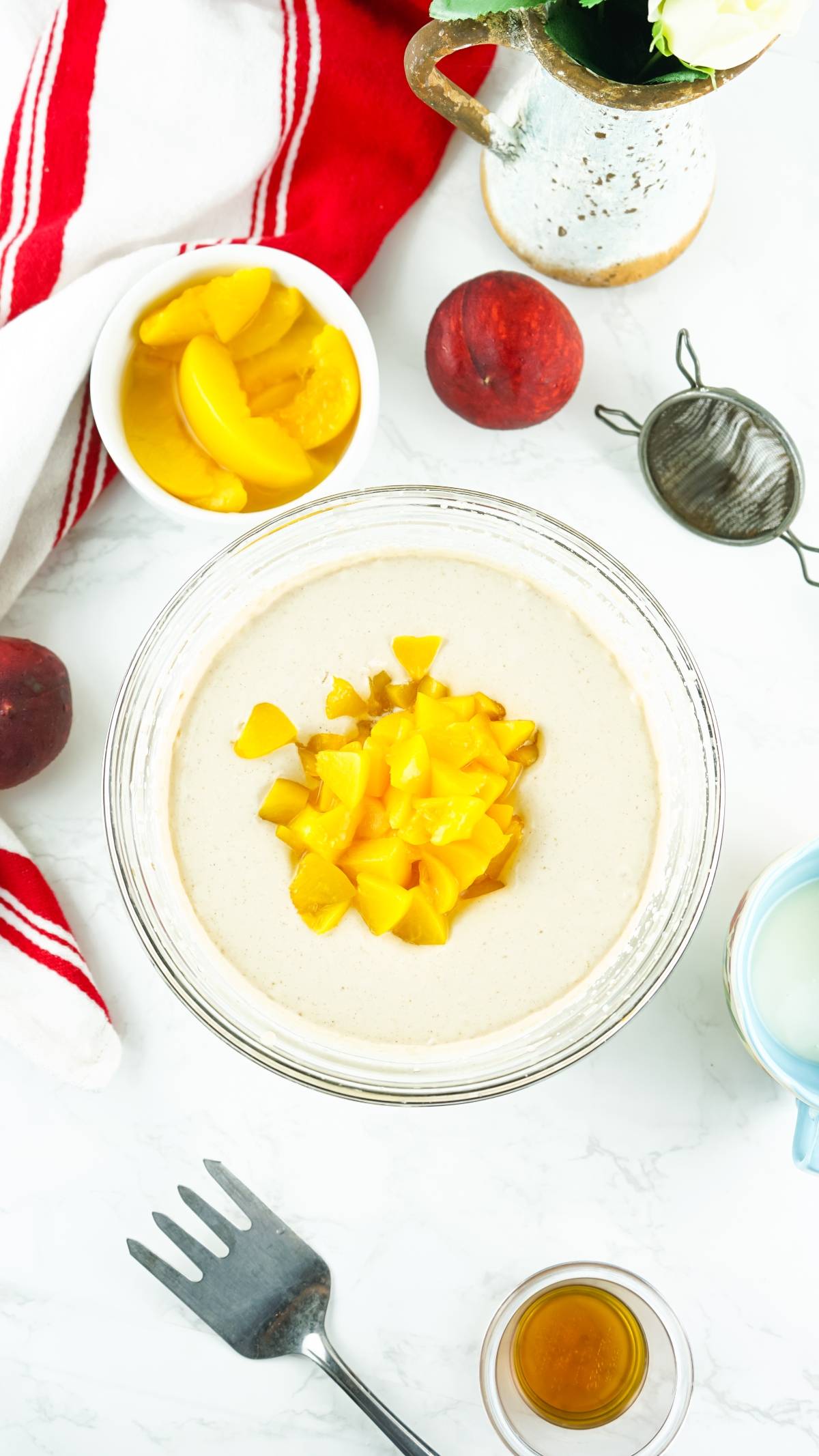 Bowl of batter topped with diced peaches, surrounded by peaches, cream, vanilla, and a red-striped towel—perfect for creating a no churn peach ice cream or a sweet boozy peach dessert made with canned peaches.