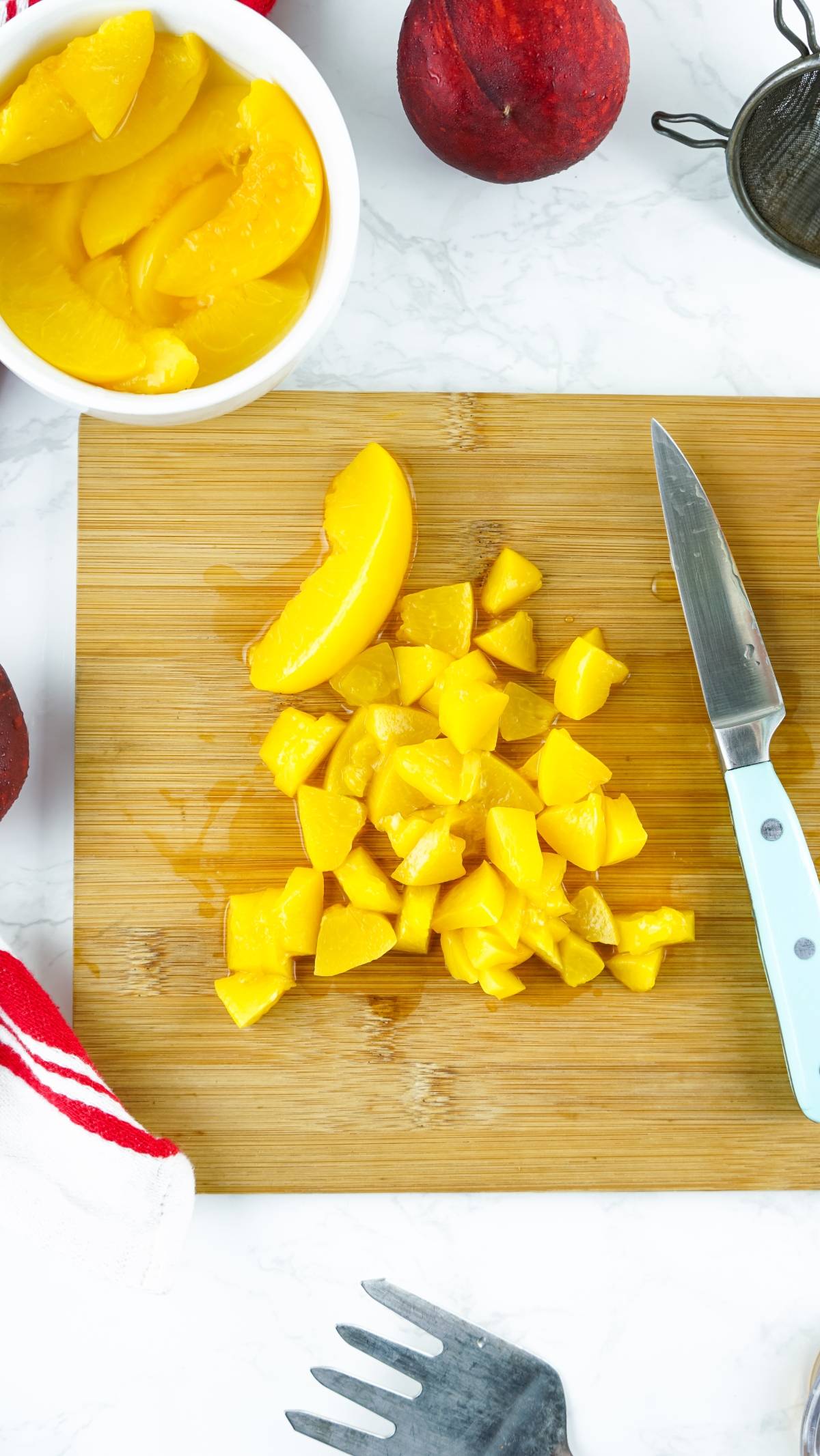 Chopped and sliced peaches on a cutting board with a knife, bowl of peaches, and peaches nearby—perfect for making no churn peach ice cream or desserts made with canned peaches.