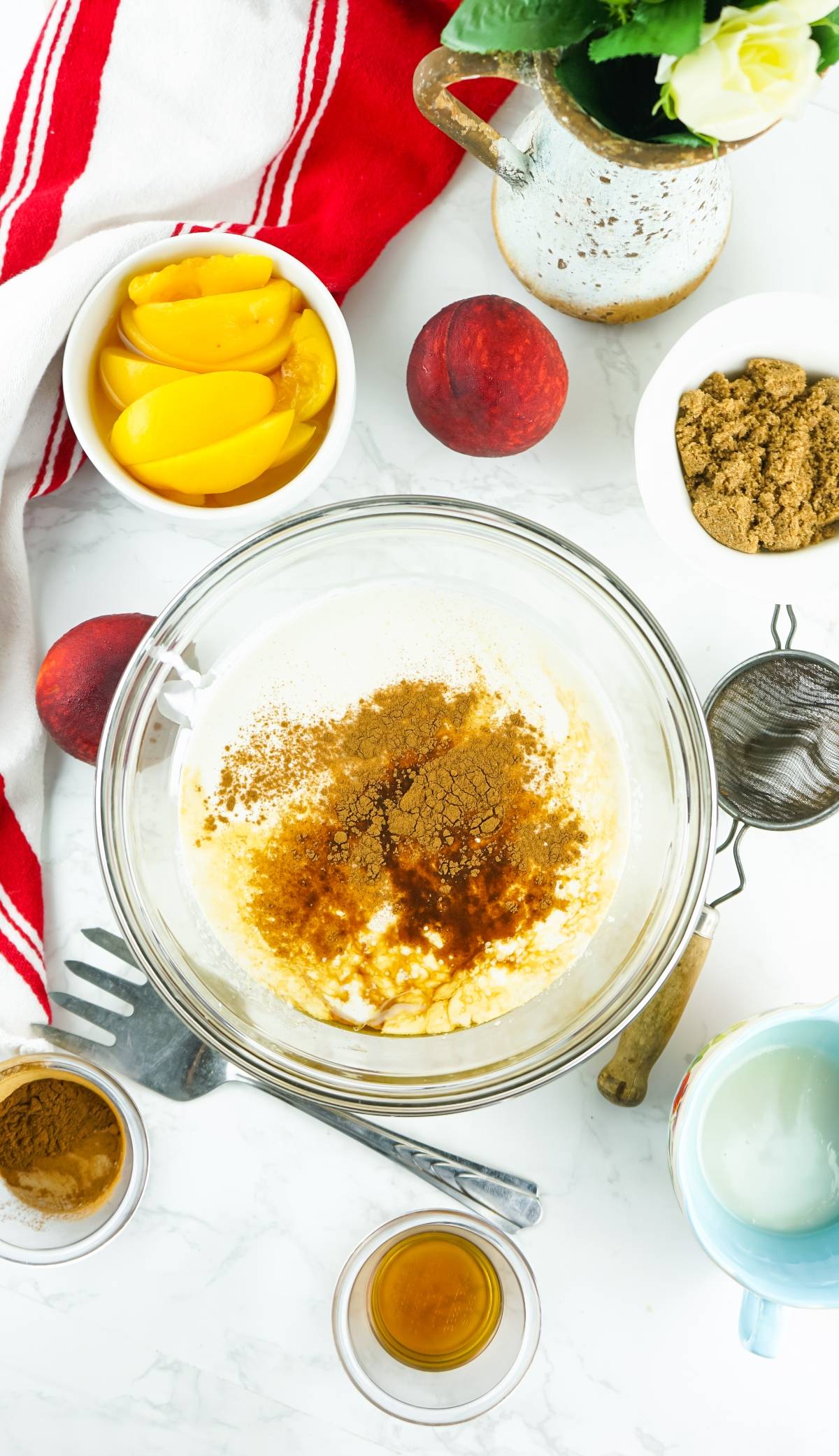 Bowl of batter with cinnamon, surrounded by canned peaches, brown sugar, vanilla, and kitchen utensils on a white table—perfect for making no churn peach ice cream.