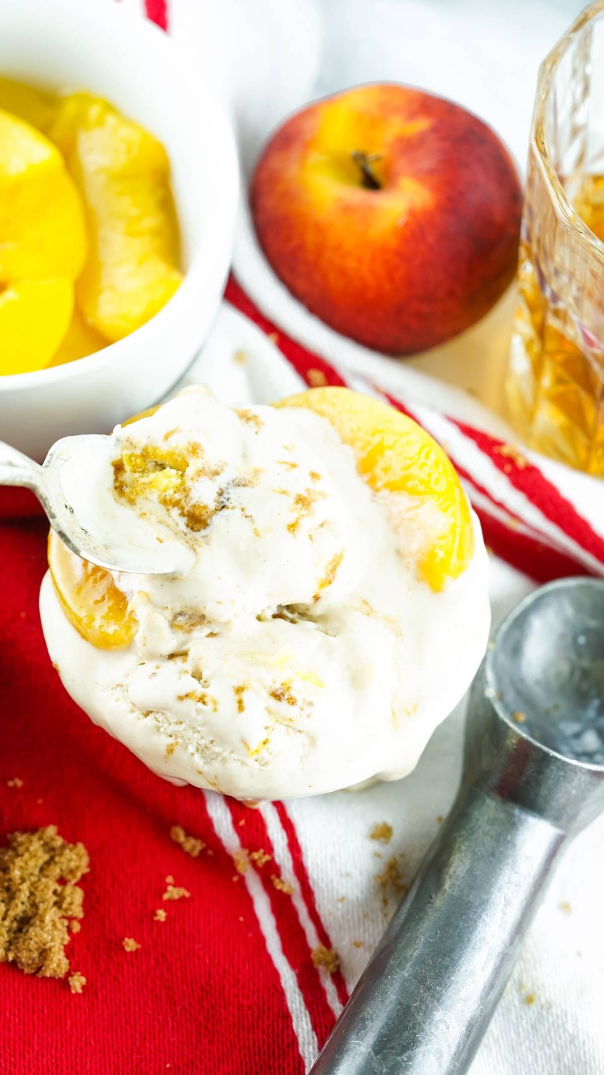 Bowl of no churn peach ice cream with peach slices and crumbled topping, next to a peach and glass on a red cloth—made with canned peaches for easy indulgence.