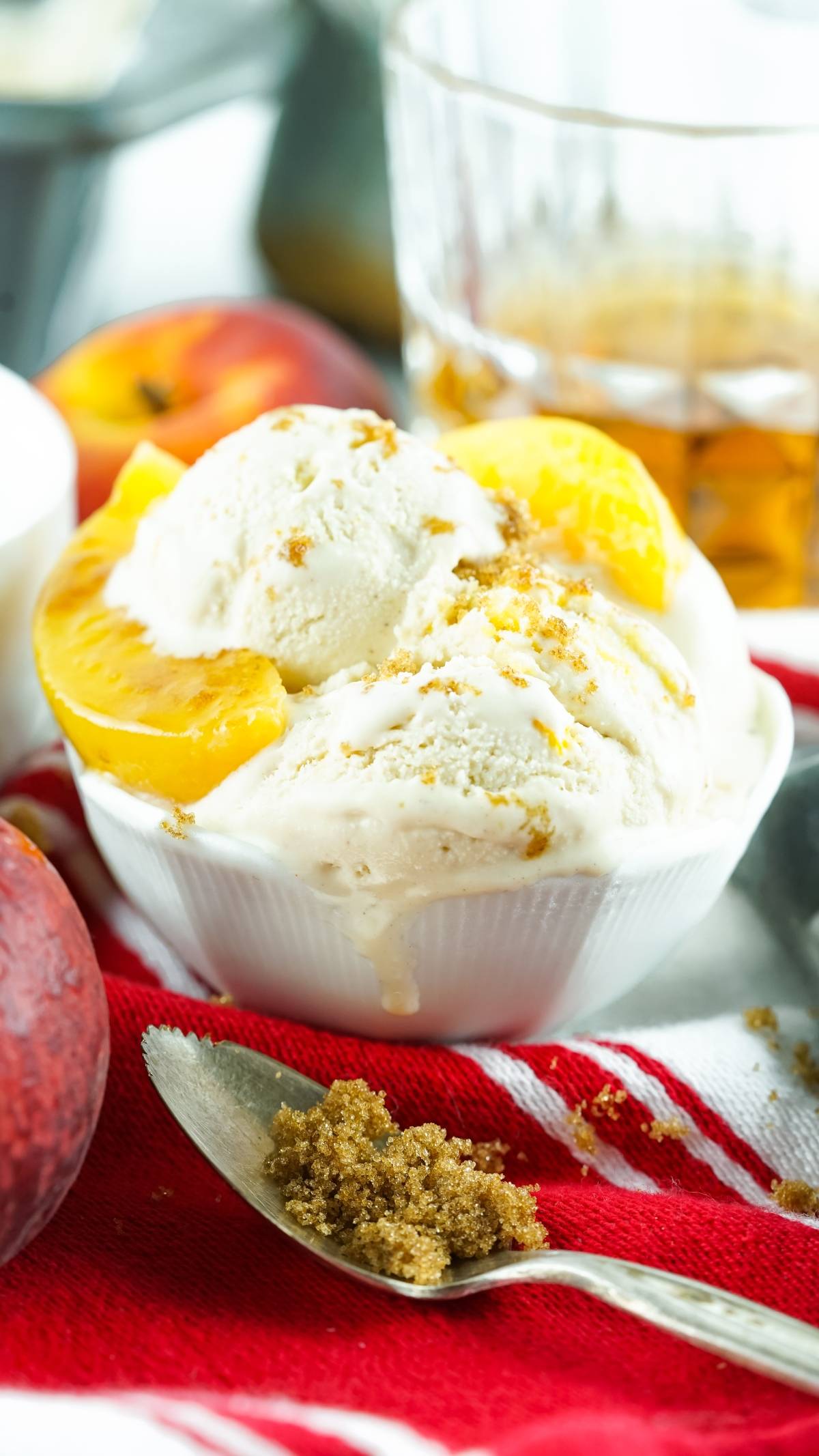 No Churn Bourbon Peach Ice Cream with peach slices and brown sugar in a white bowl on a red cloth, served with a spoon nearby.