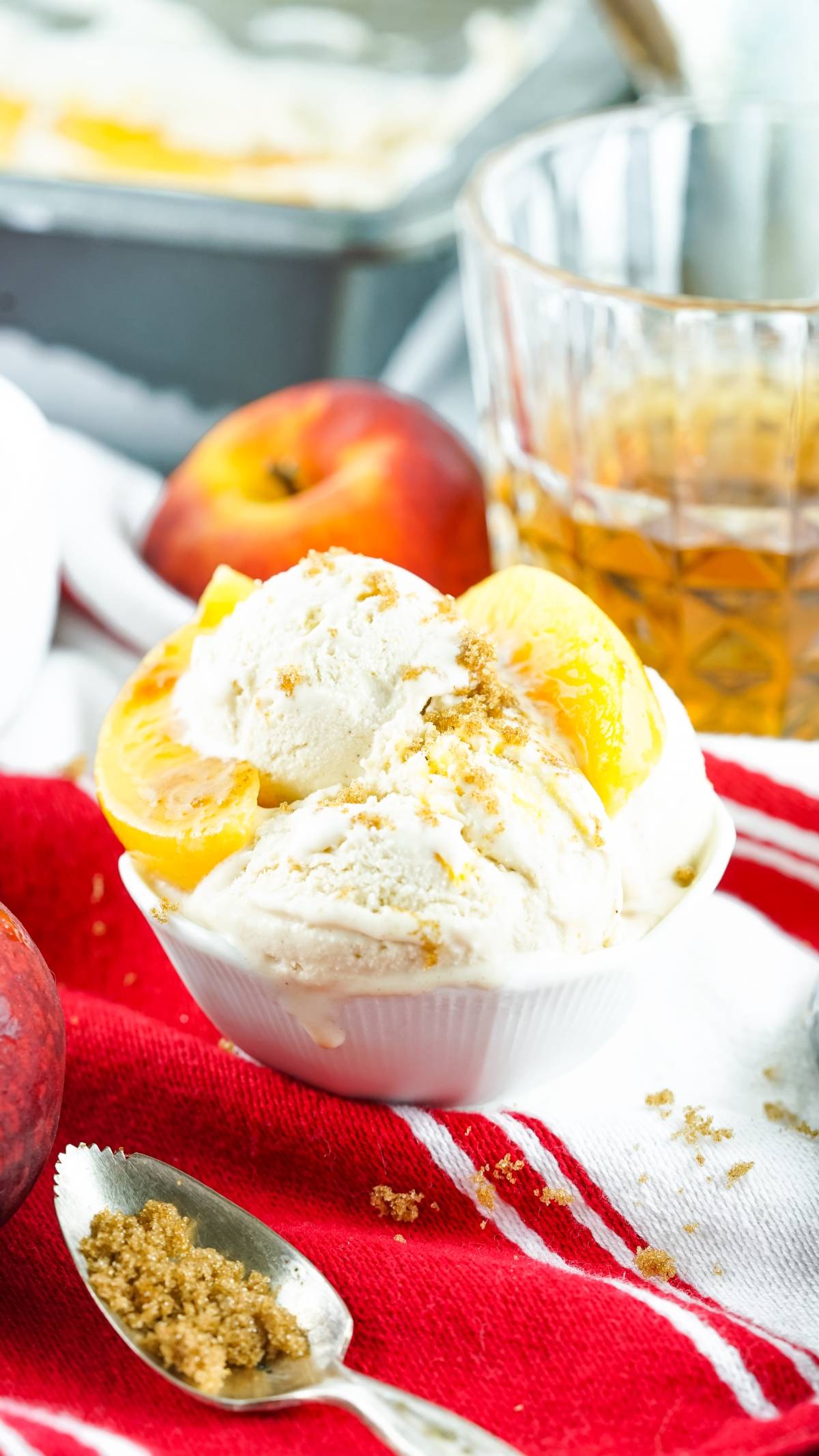 Vanilla ice cream with peach slices made with canned peaches and brown sugar in a bowl, on a red cloth, with fresh peaches and a boozy peach dessert drink nearby.