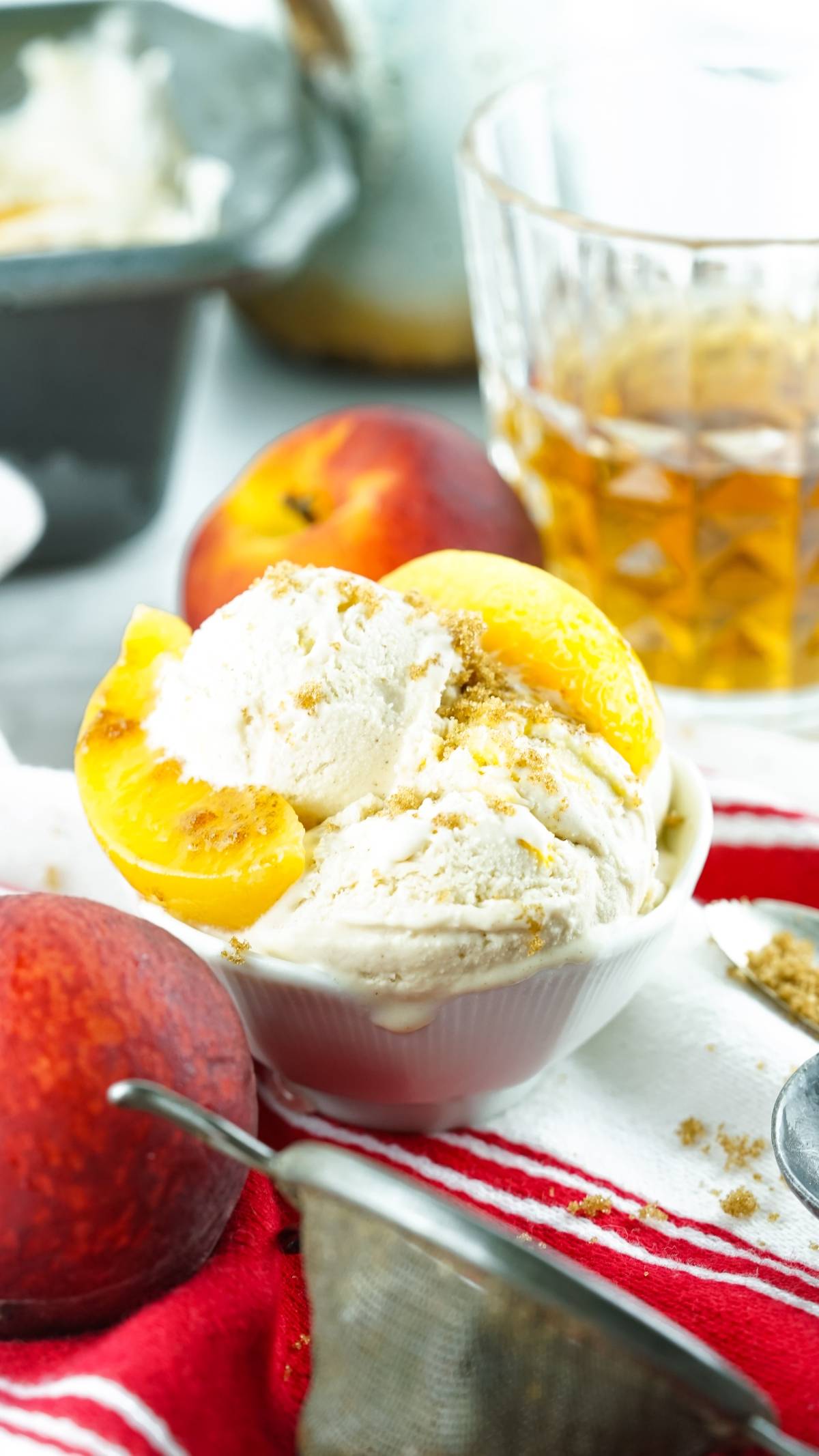 Bowl of no churn peach ice cream topped with peach slices and crumbles, next to fresh peaches and a glass of iced tea.