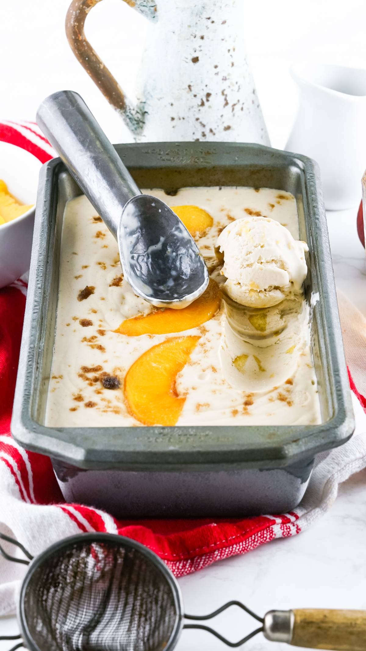 A metal pan of no churn peach ice cream, made with canned peaches, features peach slices and an ice cream scoop resting on top.