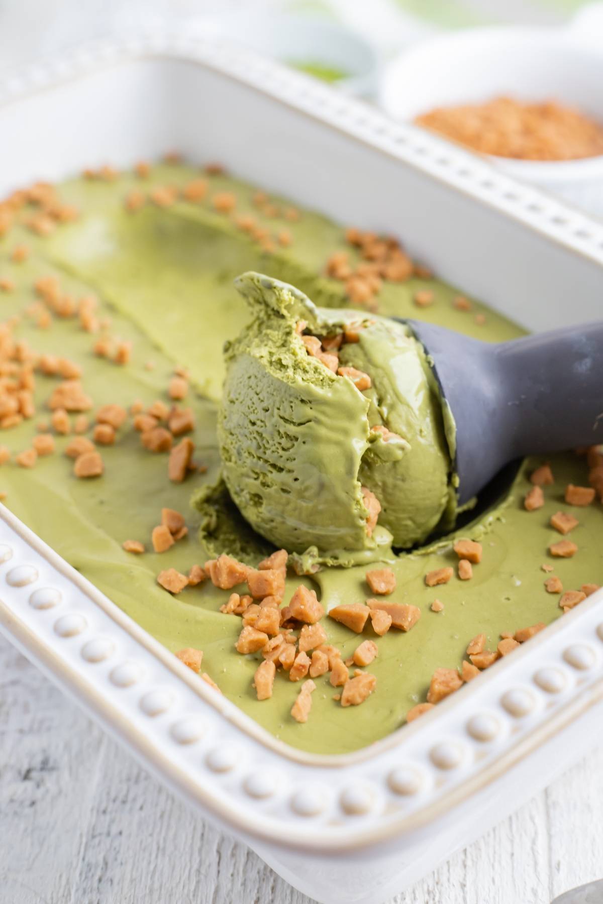 A scoop of green tea ice cream with toffee bits—this No Churn Matcha Dulce de Leche Ice Cream recipe is served in a white dish on a wooden surface.