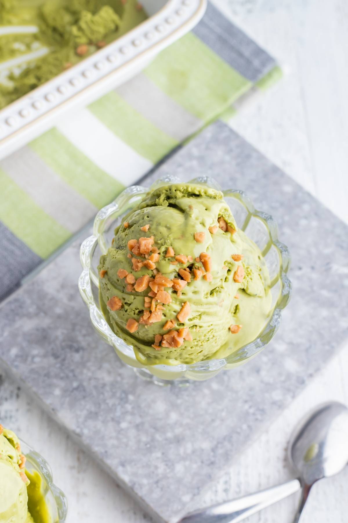 A glass dish of green tea ice cream, made with a no churn ice cream recipe, topped with crunchy bits on a gray stone slab, with a spoon nearby.