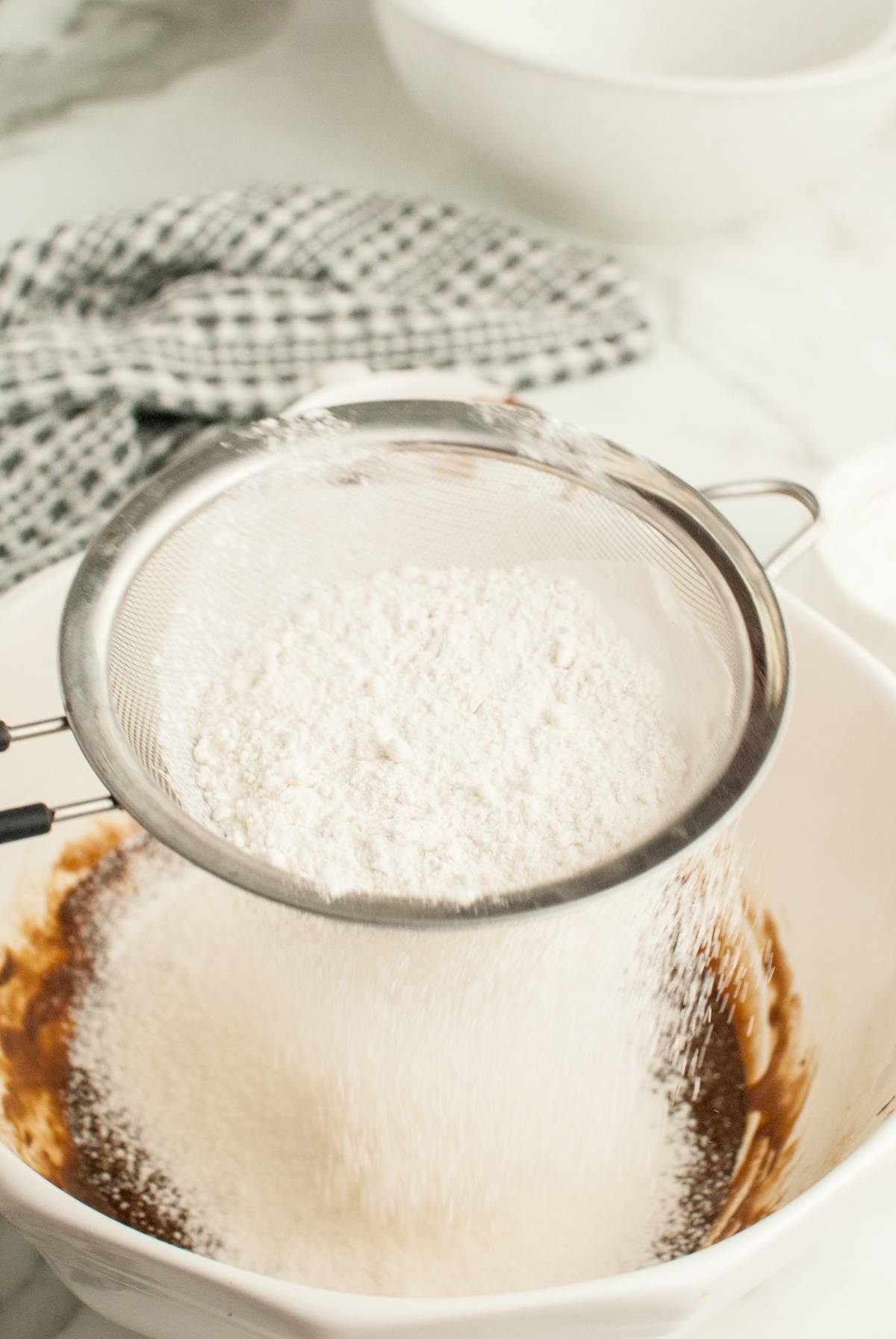 Flour being sifted into a bowl of batter, perfect for making Nutellotti - Italian Nutella Cookies, with a kitchen towel and bowl in the background.