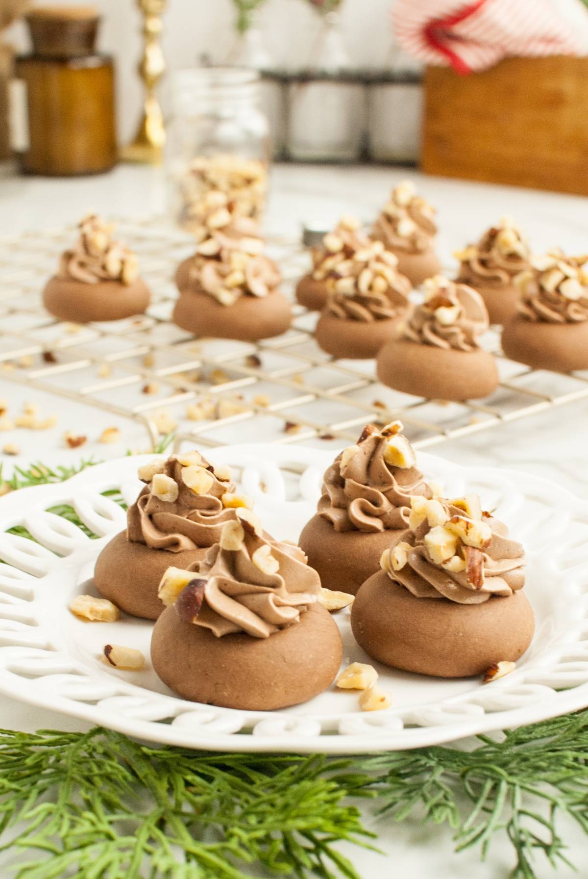 Chocolate cookies topped with swirls of chocolate frosting and chopped nuts, these Italian Nutella cookies are beautifully arranged on a white plate and cooling rack.