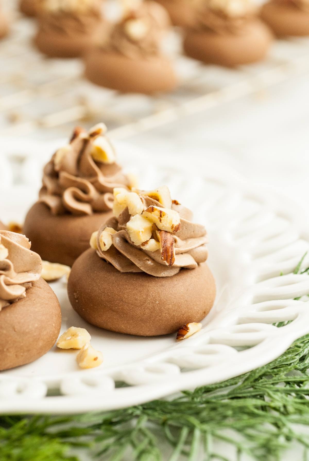 Chocolate cookies topped with swirls of chocolate frosting and chopped nuts on a white decorative plate—a festive treat inspired by Italian Nutella cookies perfect for holiday gatherings.