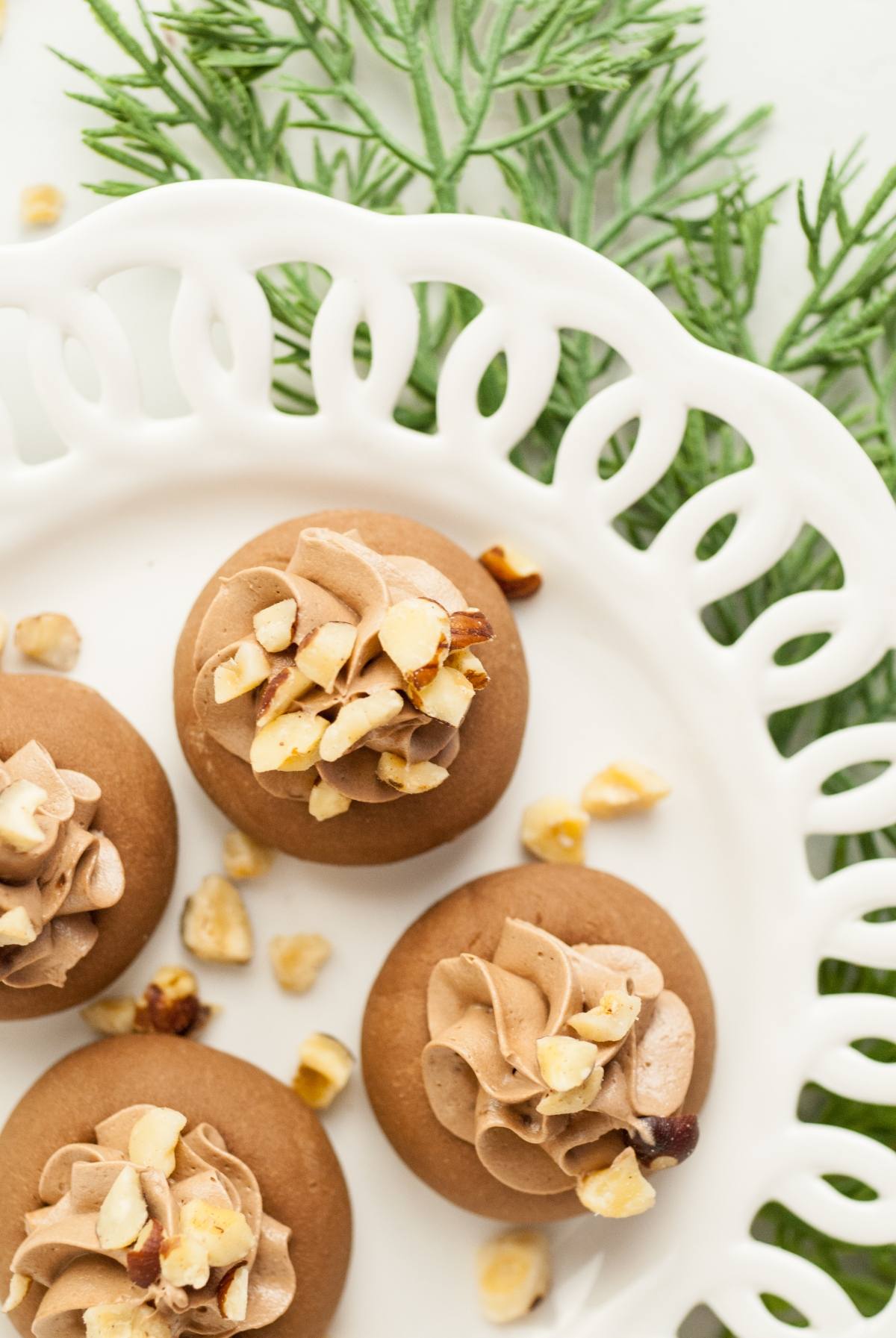 Chocolate mousse cookies topped with chopped nuts on a white plate, reminiscent of Nutella thumbprint cookies, with green foliage in the background.