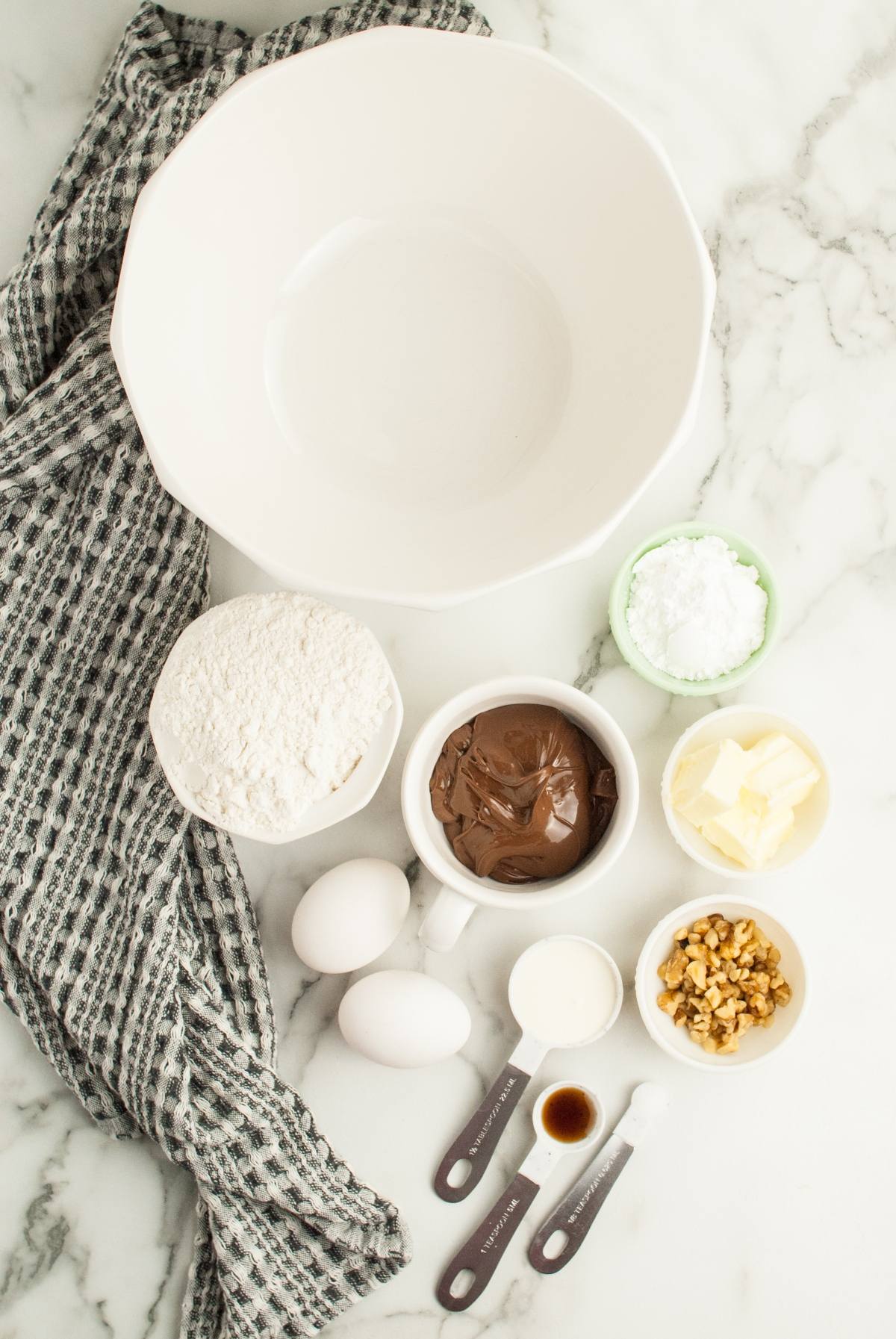 Baking ingredients in bowls with measuring spoons and a large empty bowl on a marble countertop—perfect for whipping up Nutellotti, classic Italian Nutella cookies, or exploring other recipes that use Nutella.