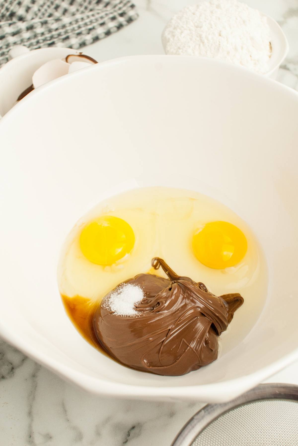 A mixing bowl with two eggs, chocolate spread, vanilla, and sugar on a marble countertop—perfect for making Nutellotti - Italian Nutella Cookies.