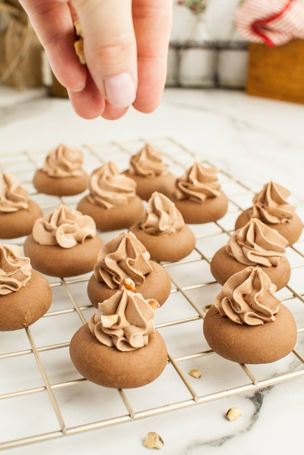 A hand sprinkles chopped nuts on Nutellotti - Italian Nutella Cookies with swirled frosting, arranged on a cooling rack.