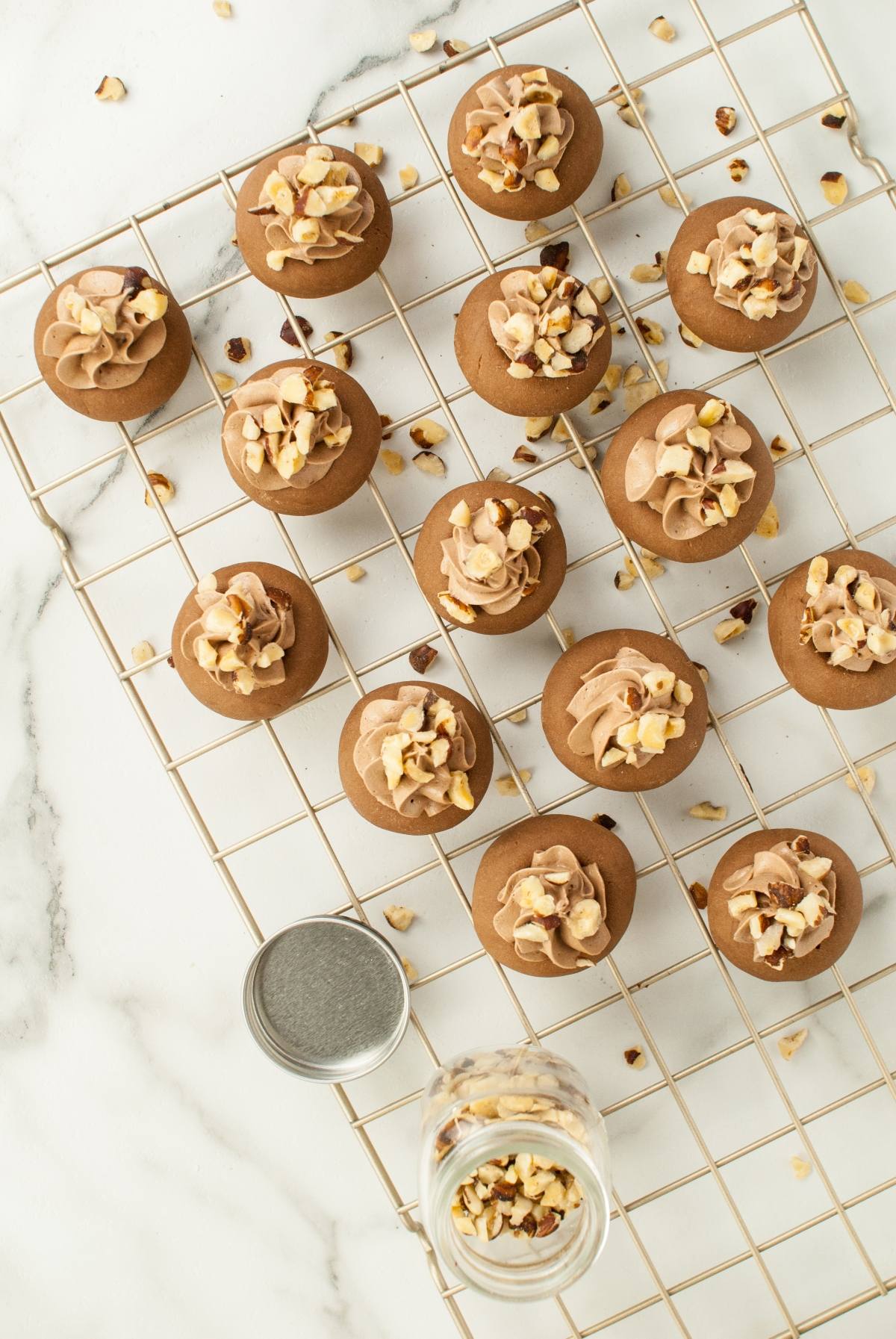 Chocolate cookies with frosting and chopped nuts on a cooling rack, inspired by classic Italian Nutella cookies. A jar of nuts sits nearby on a marble surface—perfect for anyone seeking delicious recipes that use Nutella.