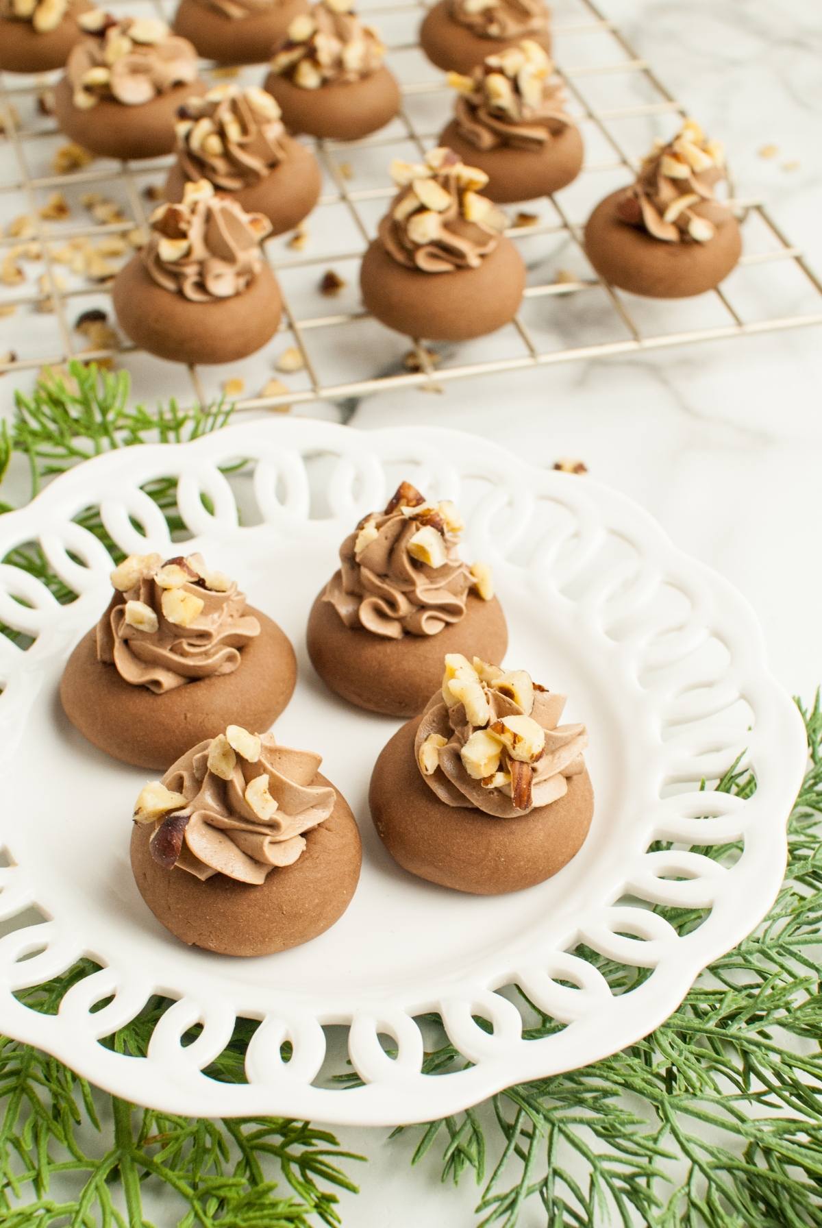 Chocolate cookies with piped frosting and chopped nuts on a white plate, with more Nutellotti – Italian Nutella Cookies cooling on a rack in the background.