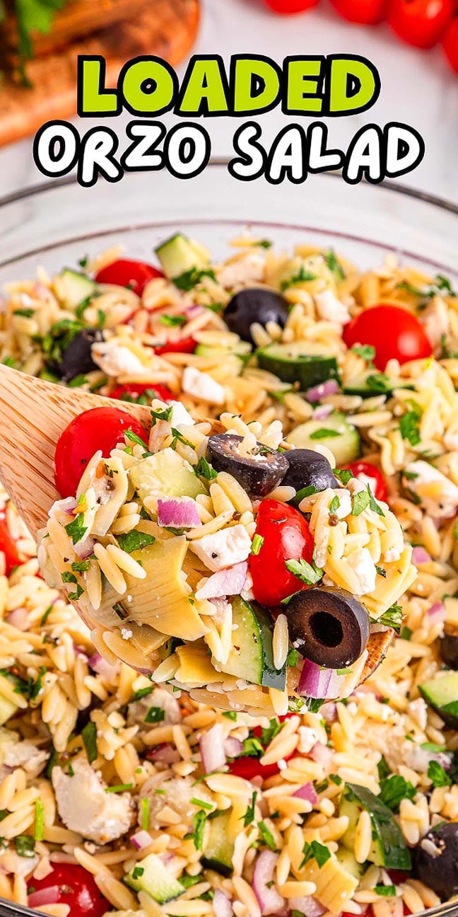 A wooden spoon holds vibrant orzo salad with tomatoes, olives, cucumber, and herbs above a large bowl.
