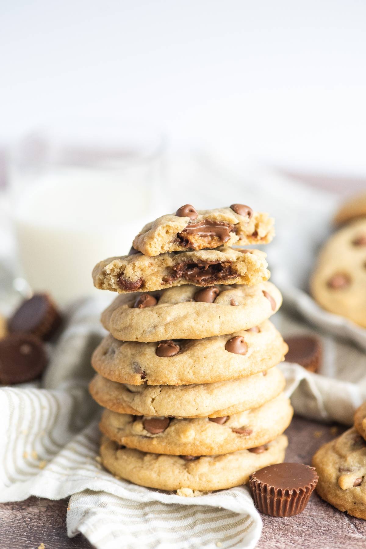 A stack of stuffed chocolate chip cookies with the top cookie broken in half, revealing a gooey Reese’s center, sits next to a glass of milk.