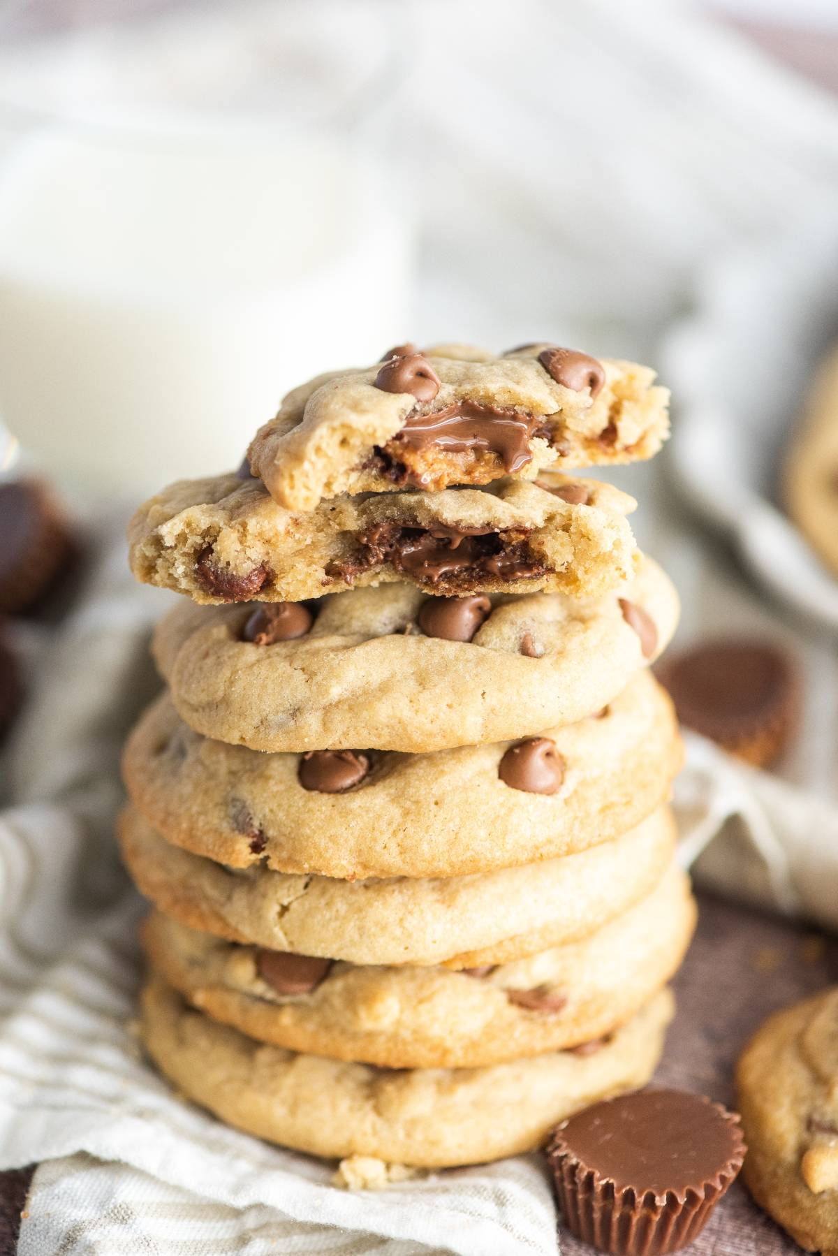A stack of peanut butter cup stuffed cookies, with the top cookie broken in half to reveal the gooey center, sits next to a glass of milk.