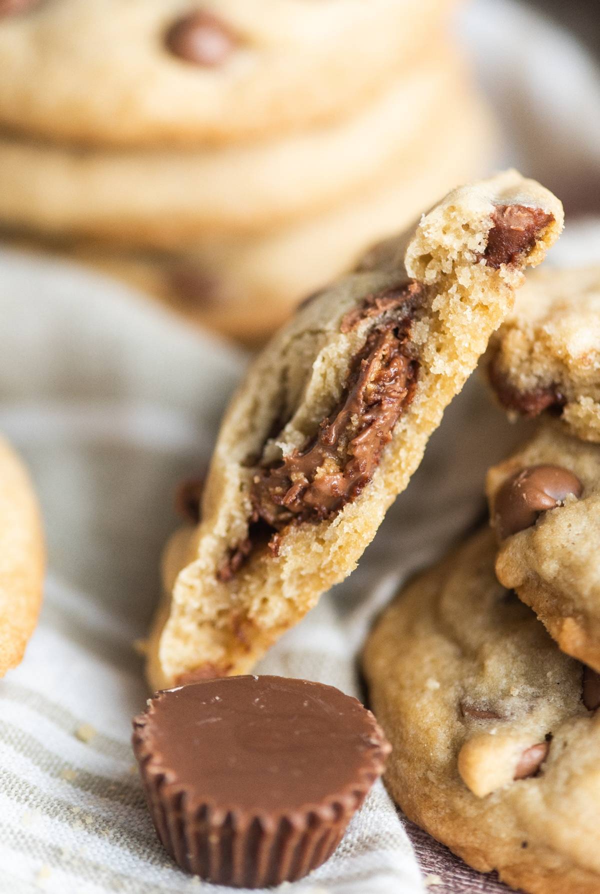 A stuffed chocolate chip cookie broken in half reveals a gooey peanut butter cup inside, next to a whole peanut butter cup.