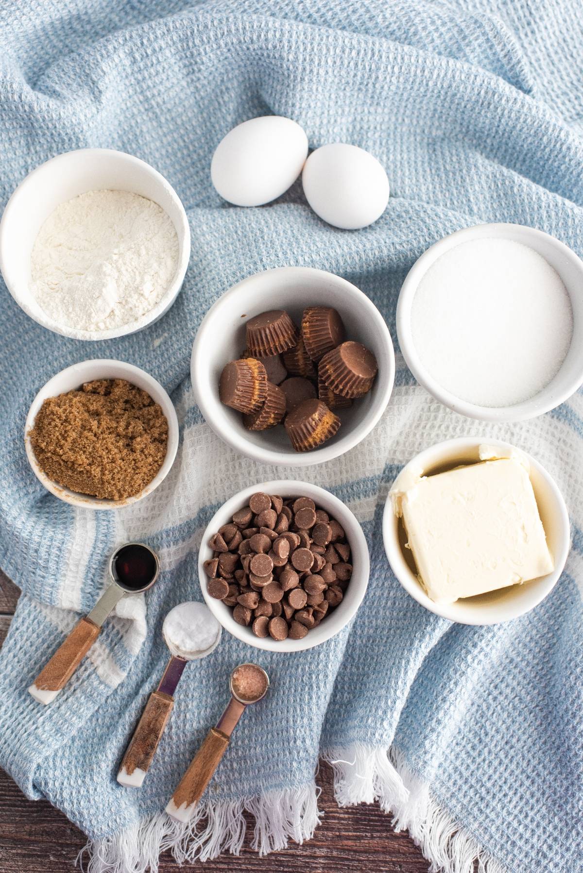 Baking ingredients in bowls: flour, sugar, eggs, butter, chocolate chips, and mini peanut butter cups—perfect for peanut butter cup stuffed cookies or cookies with Reese’s in the middle. Measuring spoons included.