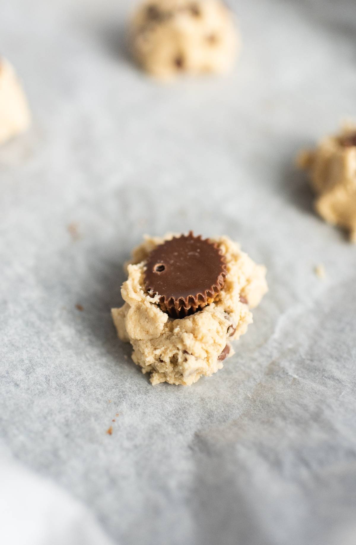 Cookie dough with a Reese’s in the middle on a sheet of parchment paper, these peanut butter cup stuffed cookies are the ultimate treat for chocolate lovers.