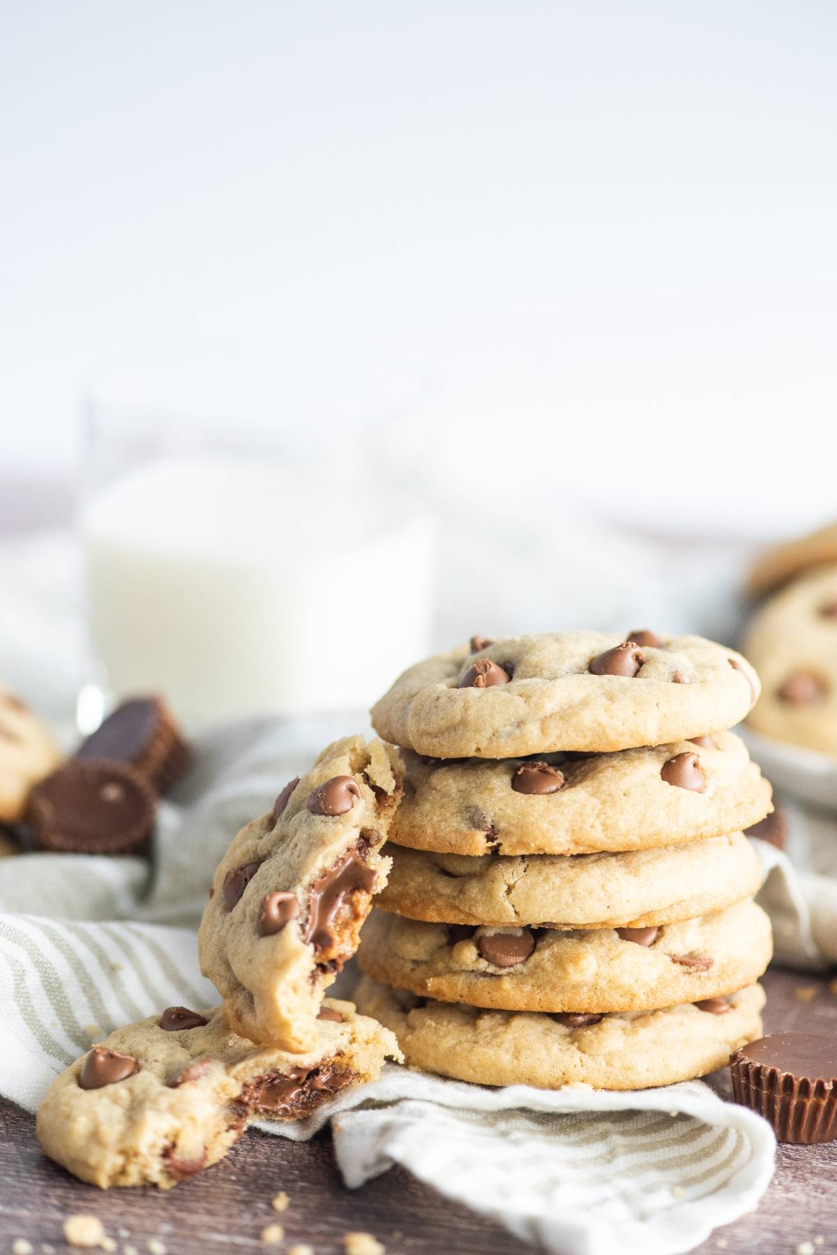 A stack of cookies with Reese’s in the middle sits next to a glass of milk; one stuffed chocolate chip cookie is broken open in front.