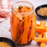 A jar of quick pickled carrots is surrounded by raw carrot sticks and small bowls of spices on a table.