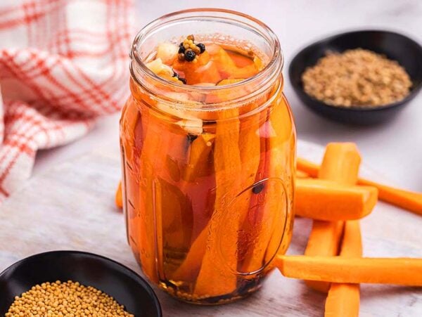 A jar of quick pickled carrots is surrounded by raw carrot sticks and small bowls of spices on a table.