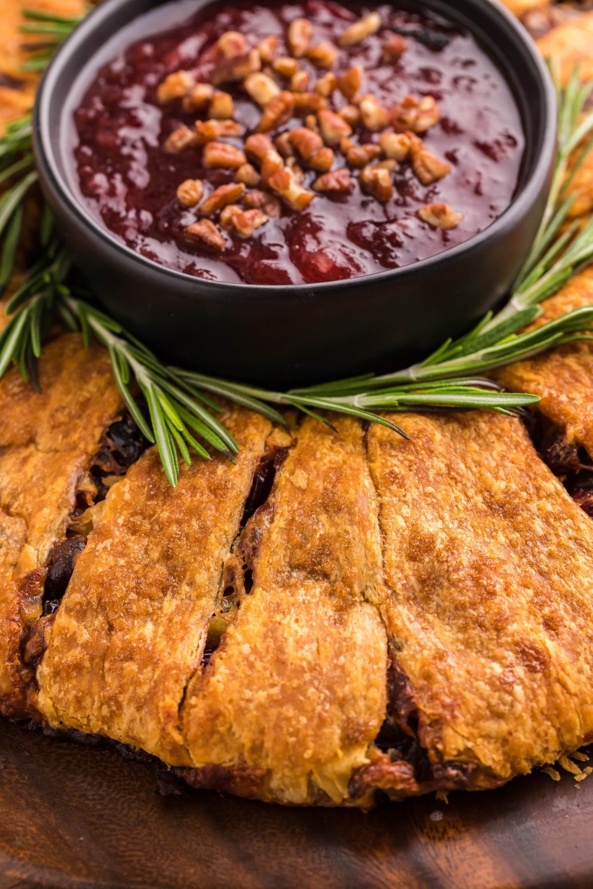 Golden baked pastry, shaped like a Christmas wreath, filled with brie and a bowl of cranberry sauce topped with pecans, garnished with fresh rosemary.