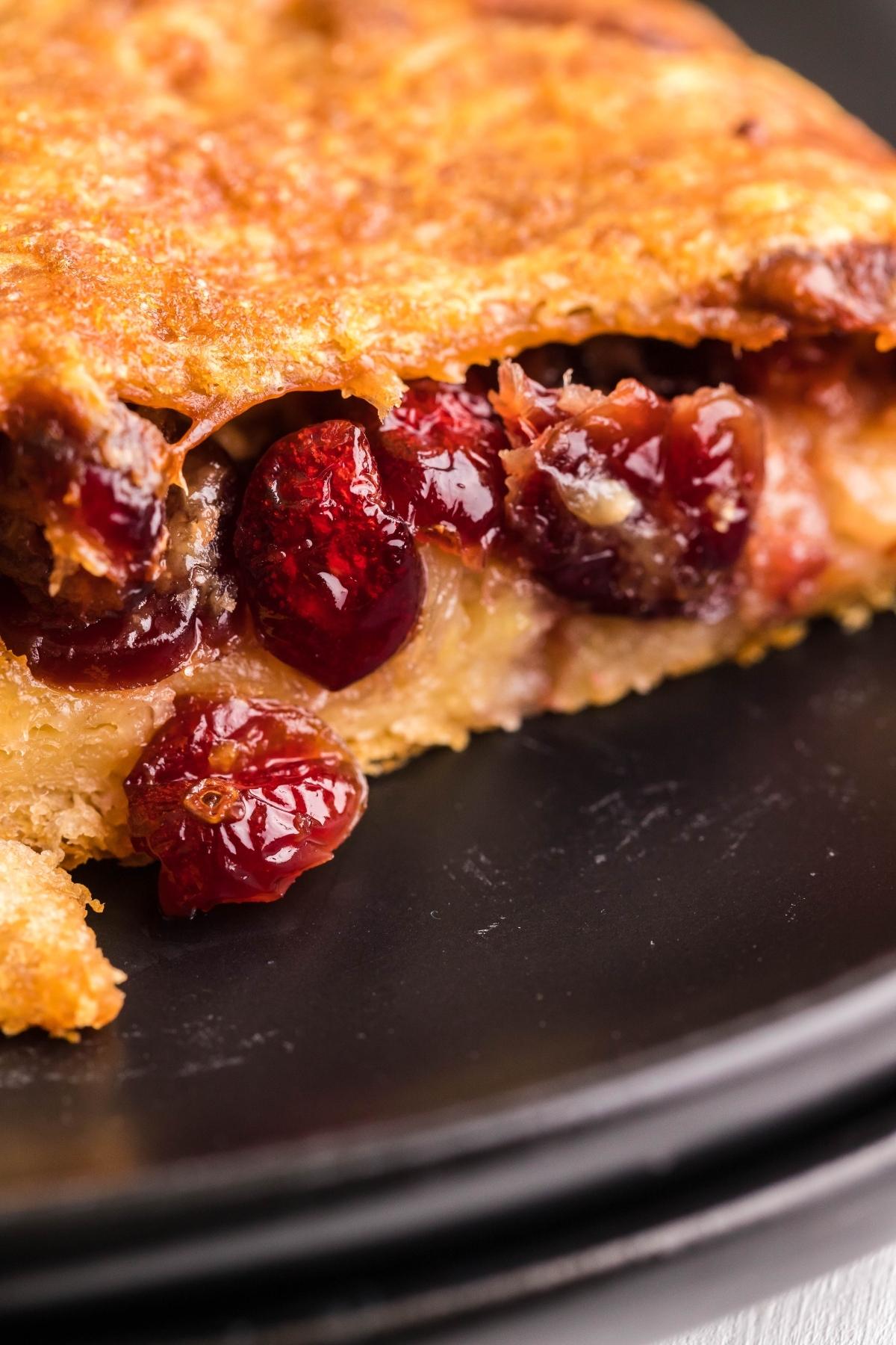 Close-up of a slice of pie with a golden crust and juicy red berries, reminiscent of a festive cranberry brie wreath, served on a black plate.