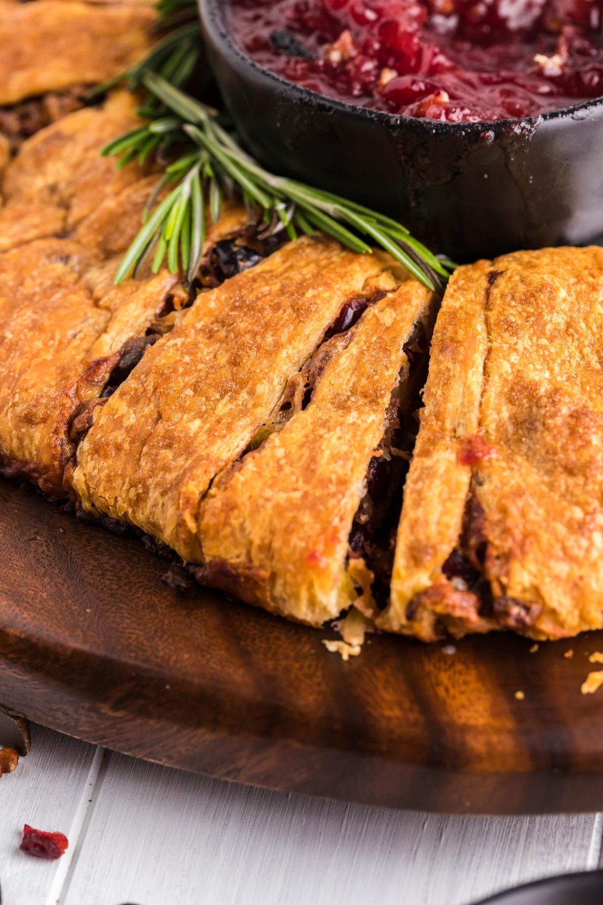 Close-up of a festive Pineapple Cranberry Brie Wreath with a bowl of red dipping sauce and sprigs of rosemary on a wooden plate, perfect for serving as a delicious Christmas wreath appetizer.